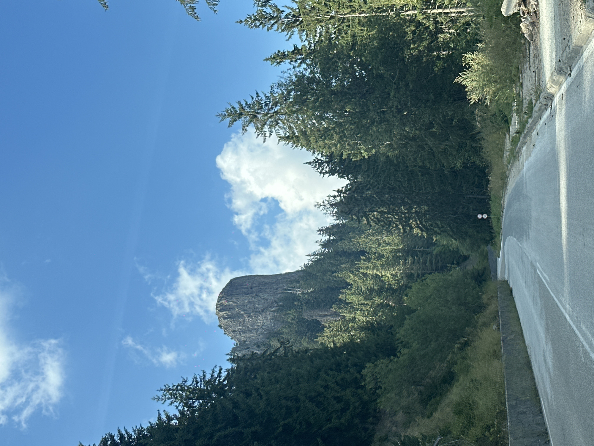 The image depicts a winding asphalt road through a mountainous, forested area under a bright blue sky with scattered white clouds. The road curves to the right, bordered by a concrete barrier on the left and dense coniferous trees on the right. In the background, a prominent, sheer rock formation rises above the treeline. The scene appears to be located in the area of Smolyan, Bulgaria, known for its Rhodope Mountains scenery. There are no people or overt actions visible in the image, with the focus being on the natural landscape and the road infrastructure.