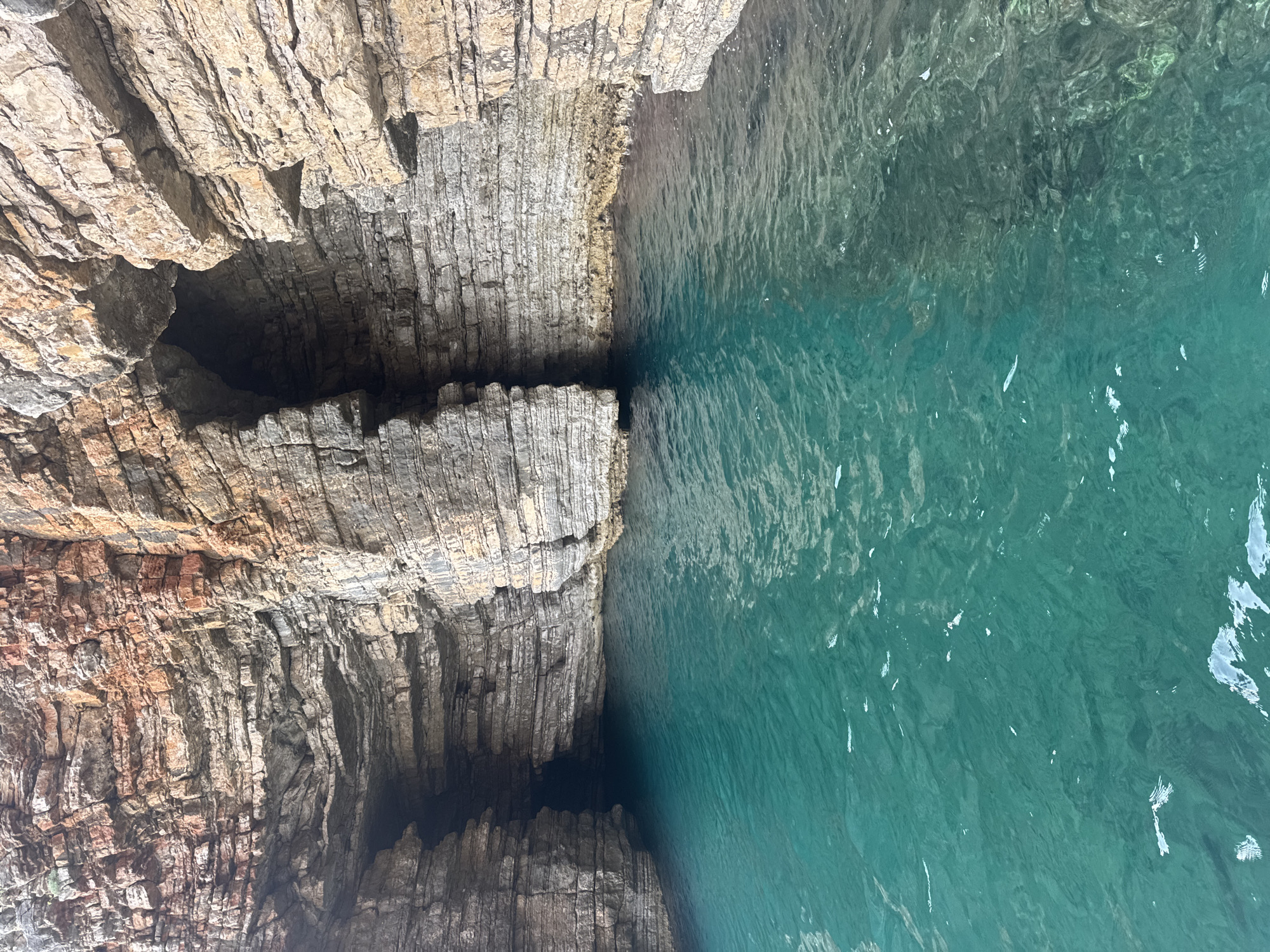 The image shows a view of the sea cave known as the Blue Cave, located off the coast of Budva, Montenegro. The cave entrance is visible on the right side of the image, with turquoise water filling the foreground. The cave walls are made of layered rock formations, with some areas appearing dark and others illuminated by sunlight. The water is clear and calm, with gentle ripples on the surface. The overall scene is one of natural beauty and tranquility.