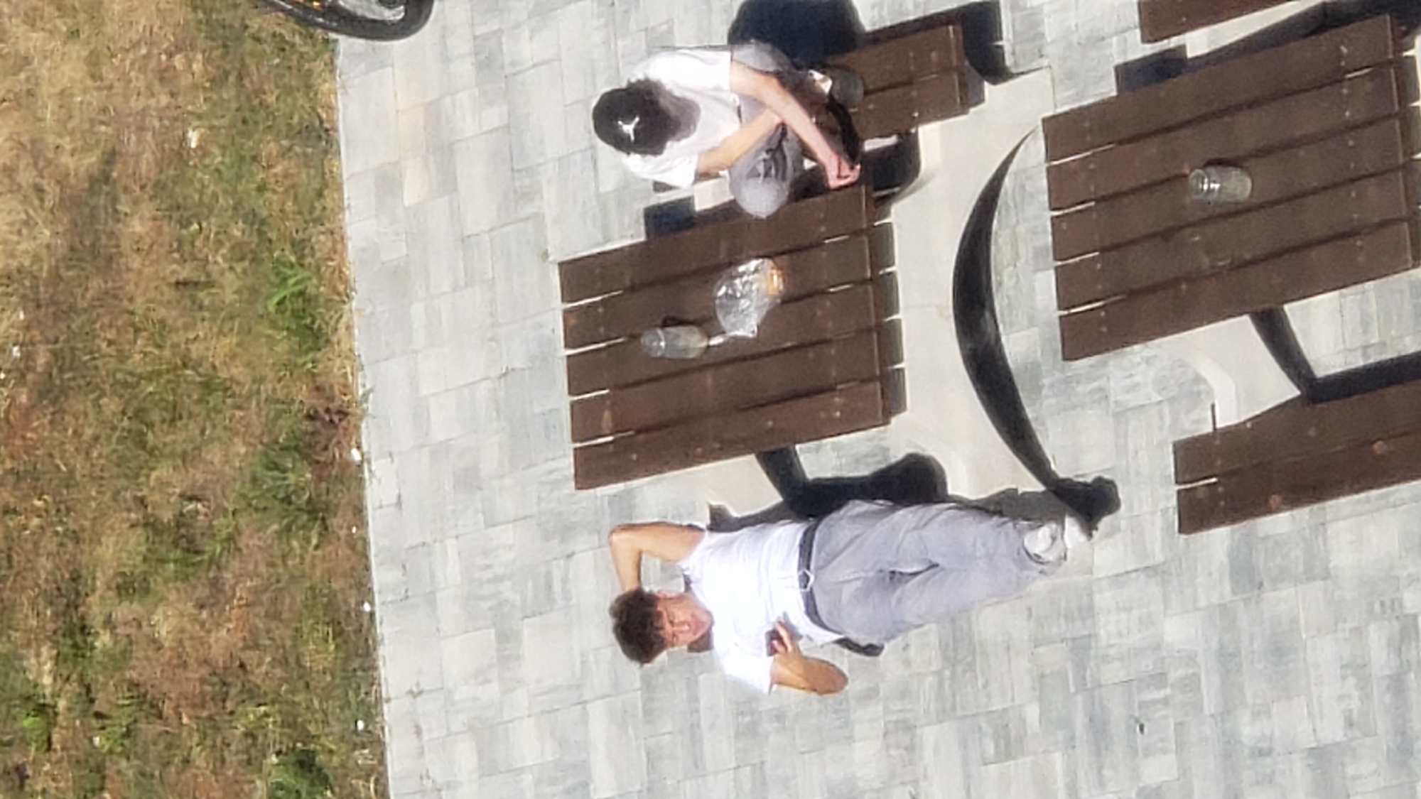 The image depicts two young males at a picnic table outdoors. One male, wearing a white t-shirt and light grey pants, is lying down with his head propped up by his hand, looking at his phone. He is positioned on the left side of the image. The other male, wearing a white t-shirt, light grey pants, and a black baseball cap with a white logo, is sitting on a bench at the picnic table, facing towards the right side of the image. He is crouched down, with his hands resting on his knees.
On the wooden picnic table, there is a glass jar and a plastic bag containing food items. The ground beneath them is paved with light-colored square tiles, and there is a patch of dry, brown grass with some green vegetation in the background. A black bicycle wheel is visible in the upper right corner of the image. The scene appears to be a public outdoor recreational area. The context of Prešov, Slovakia, suggests this location could be a park, a rest stop along a pathway, or a common area in a residential or commercial zone.