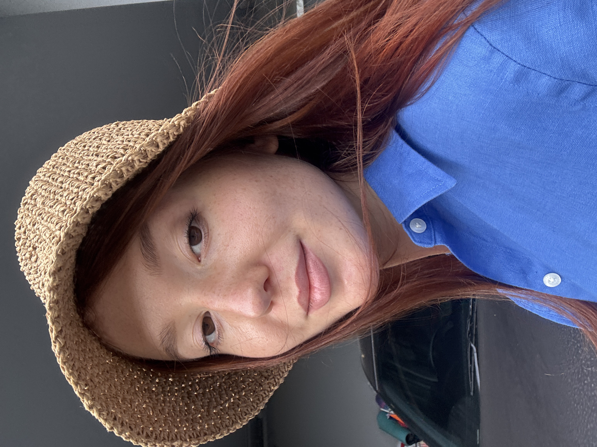 A fair-skinned person with red hair and freckles is wearing a straw hat and a blue button-down shirt. The person is facing the camera and appears to be in an indoor setting. The location in Choeng Thale, Thailand is not discernible from the image.