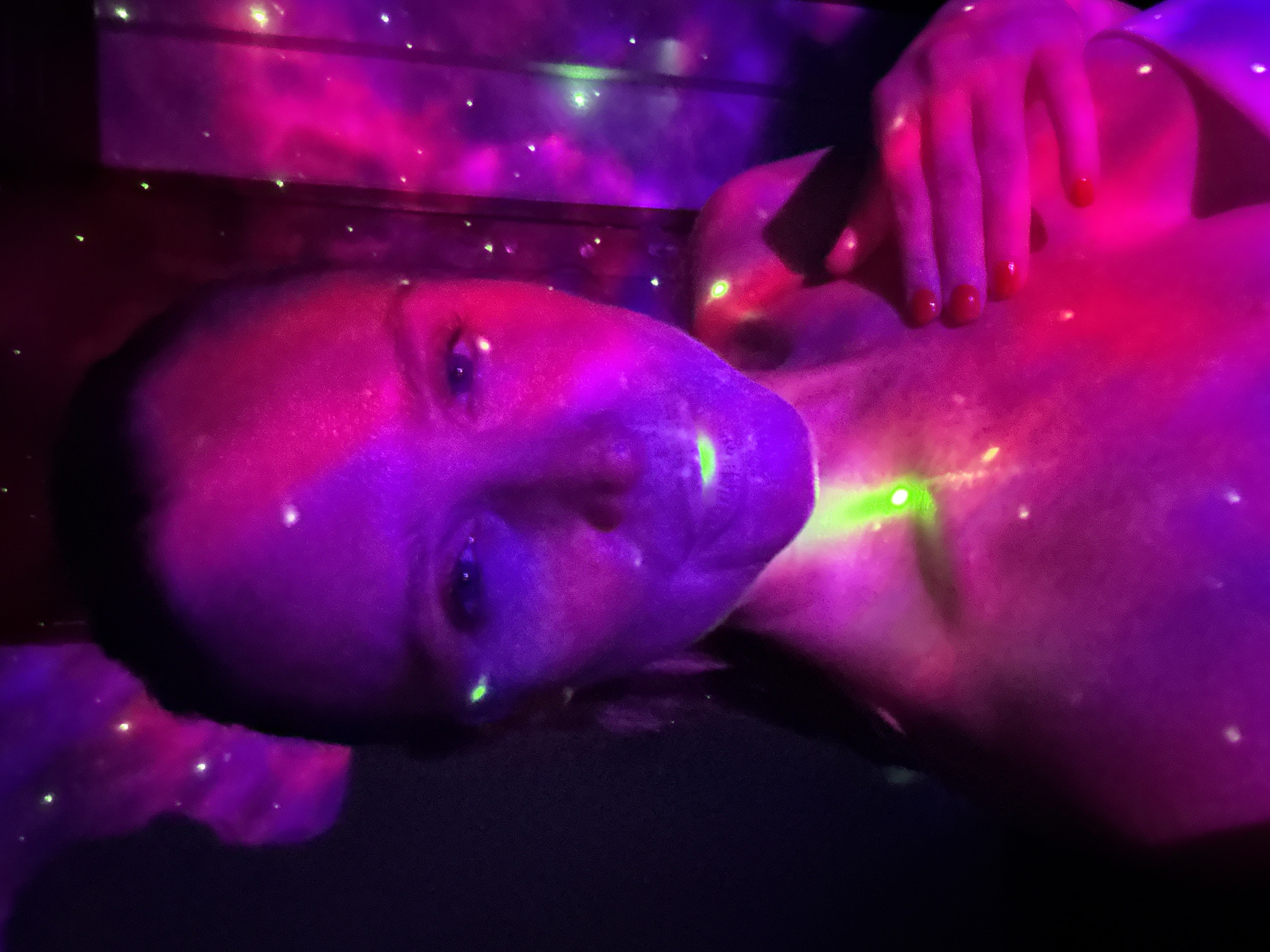 The image depicts a person, likely an adult female, lying down in a dimly lit environment, possibly an indoor space. Her head is turned towards the camera, with her eyes open, and her face is illuminated by vibrant, artificial purple and pink light. A prominent green laser dot rests on her chest, while numerous smaller green and white light specks are scattered across her skin and the dark background, resembling stars or glitter.
Her hand, with red nail polish, is visible resting on her chest, also bathed in the colorful light. The background features blurred areas of pink and purple light, creating a cosmic or nebula-like effect, suggesting the use of a light projector or similar device to create a starry or galaxy-like ambiance. The scene is dark, with the artificial lights providing the primary illumination, indicating it is likely nighttime or a blacked-out room. The person appears to be in a relaxed or contemplative state. No text is visible in the image. This scene is set in Kuta Selatan, Indonesia.