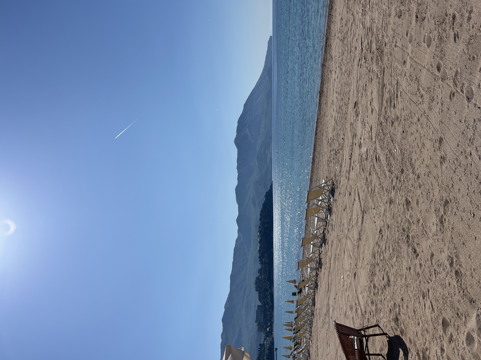 A landscape photograph captures a serene beach scene in Budva, Montenegro, on a bright, sunny day. The sky, clear and blue, dominates the upper portion of the image, with the sun creating a bright flare in the upper left corner. A faint white contrail from an airplane stretches diagonally across the upper right sky.
The deep blue sea occupies the central and mid-right sections, its surface sparkling intensely with reflected sunlight, indicating calm waters. A line of small, light-colored buoys or markers is visible in the water, extending into the distance parallel to the shore.
To the right, a sandy beach stretches out, showing numerous footprints and some textured patterns on its surface. Along the water's edge, a long row of empty, light-colored sun loungers with metal frames is neatly arranged, awaiting occupants. Closer to the foreground on the sandy beach, a single darker-colored beach chair with a woven design casts a prominent shadow to its right.
In the distance, across the calm waters, a majestic range of dark mountains rises prominently, forming the left half of the background. Some buildings and lush greenery are visible at the base of these mountains. The overall impression is one of tranquility and anticipation for leisure activities under the warm sun. There is no visible text in the image.
