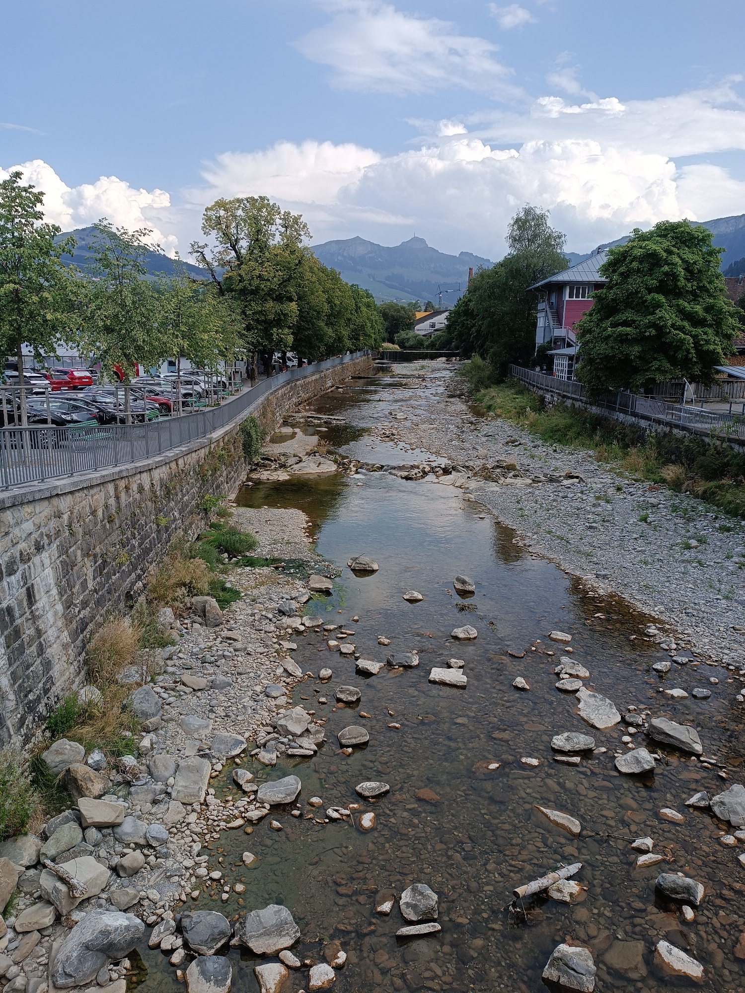 The image depicts a view of the Sitter river flowing through the town of Appenzell, Switzerland. The riverbed is lined with a variety of rocks and stones, with shallow, clear water reflecting the sky. On the left side of the river, a stone embankment supports a sidewalk and a parking lot filled with cars. Trees line the sidewalk, separating it from the parking area. Beyond the trees, buildings with red and grey facades are visible. On the right side of the river, another embankment is covered with smaller stones and grasses, with a wooden fence and walkway running along its top. Further in the distance, lush green hills and mountains rise under a sky dotted with white, puffy clouds.