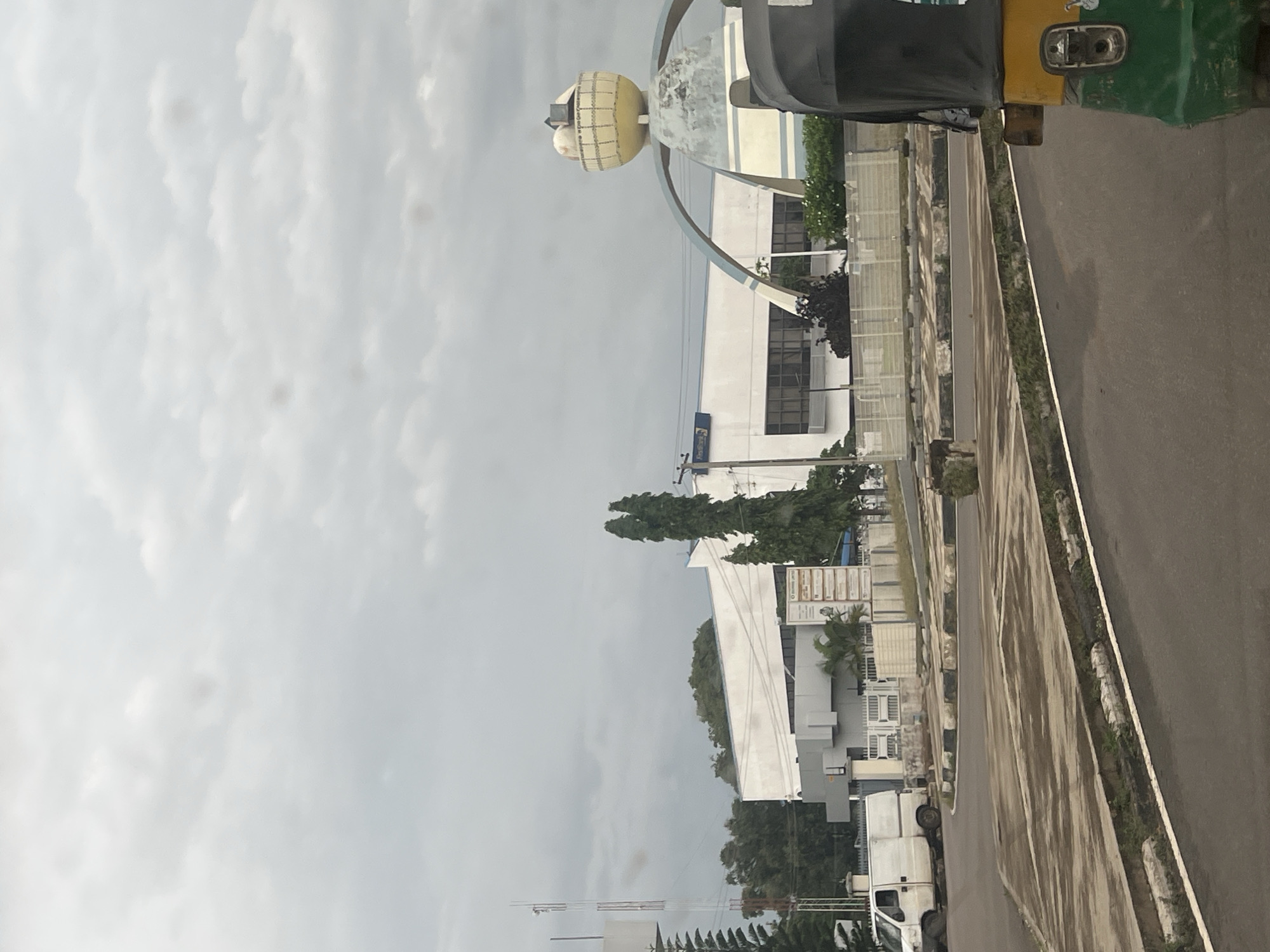 The image shows a street scene in Jos, Nigeria, on an overcast day. In the foreground, a dark-colored tuk-tuk is partially visible on the right side of the frame. The road in the foreground is paved and curves to the left. A concrete median with decorative black and white curbing separates the road from a paved area in the middle ground.
In the middle ground, a white van is parked on the left side of the frame, facing left. Beyond the van, a white building with a flat roof and dark-tinted windows is visible, appearing to be an office building or a bank. Signs are mounted on the building's facade. A tall communication tower stands to the left of the building.
A distinctive circular monument with an arch is situated to the right of the building, featuring a yellow, basket-like structure at the top, adorned with various objects. Green trees and foliage are present throughout the scene, with two tall, slender evergreen trees standing prominently near the monument and the building. The sky is overcast with diffused light.