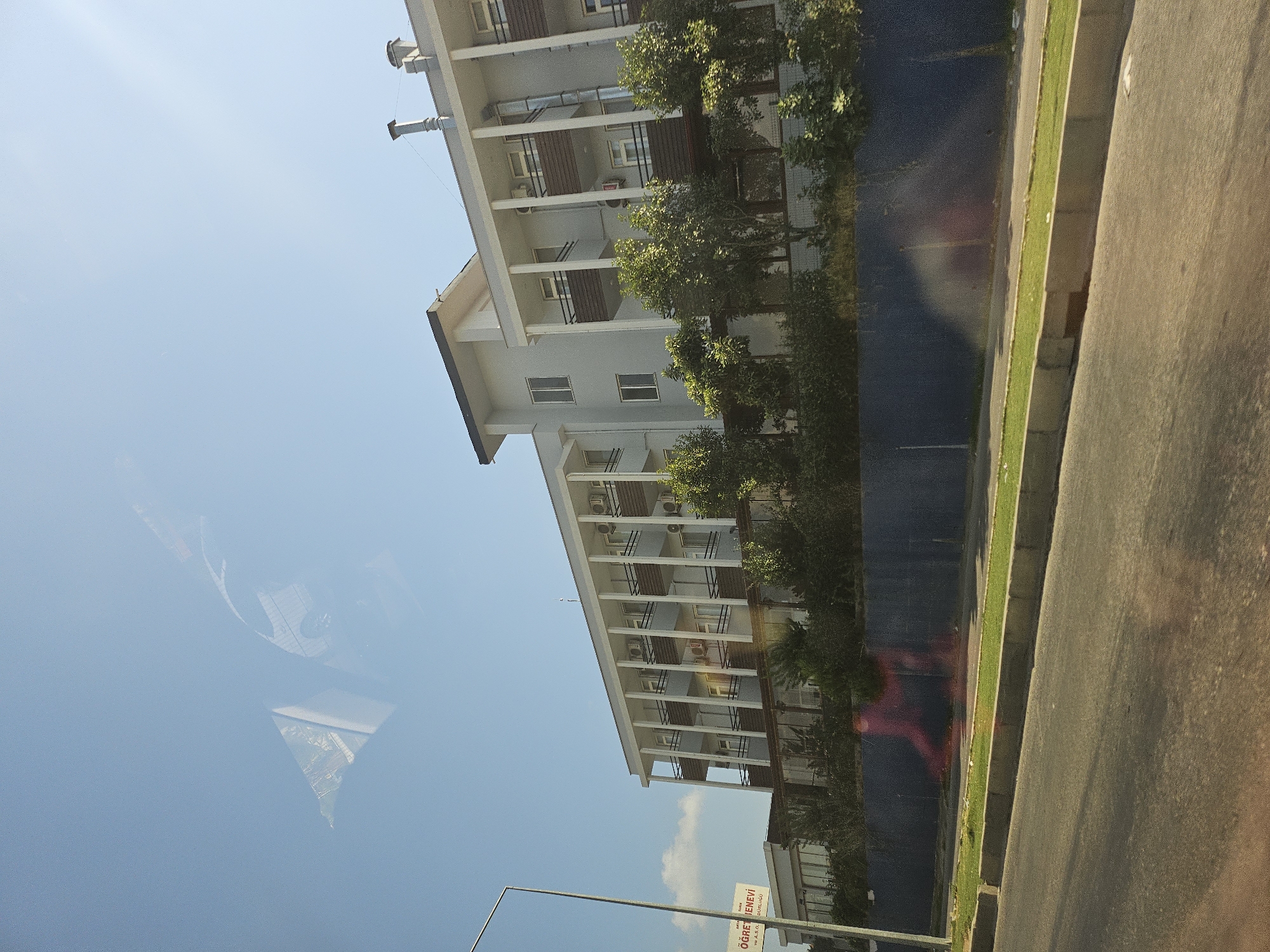 An eye-level, exterior shot captures a multi-story, light gray building with a modern architectural style. The building has a long, rectangular shape and features multiple floors with balconies and windows. The balconies are covered and have dark brown railings. The building is flanked by lush green trees. A dark blue fence runs along the front of the property, separating it from a paved road and a grassy median. A sign on a lamppost on the left side of the image reads "TÜRKİYE ÖĞRETMEN EVİ". The sky is a clear, light blue with some faint reflections visible on the surface, possibly from glass. The overall scene suggests a sunny day.