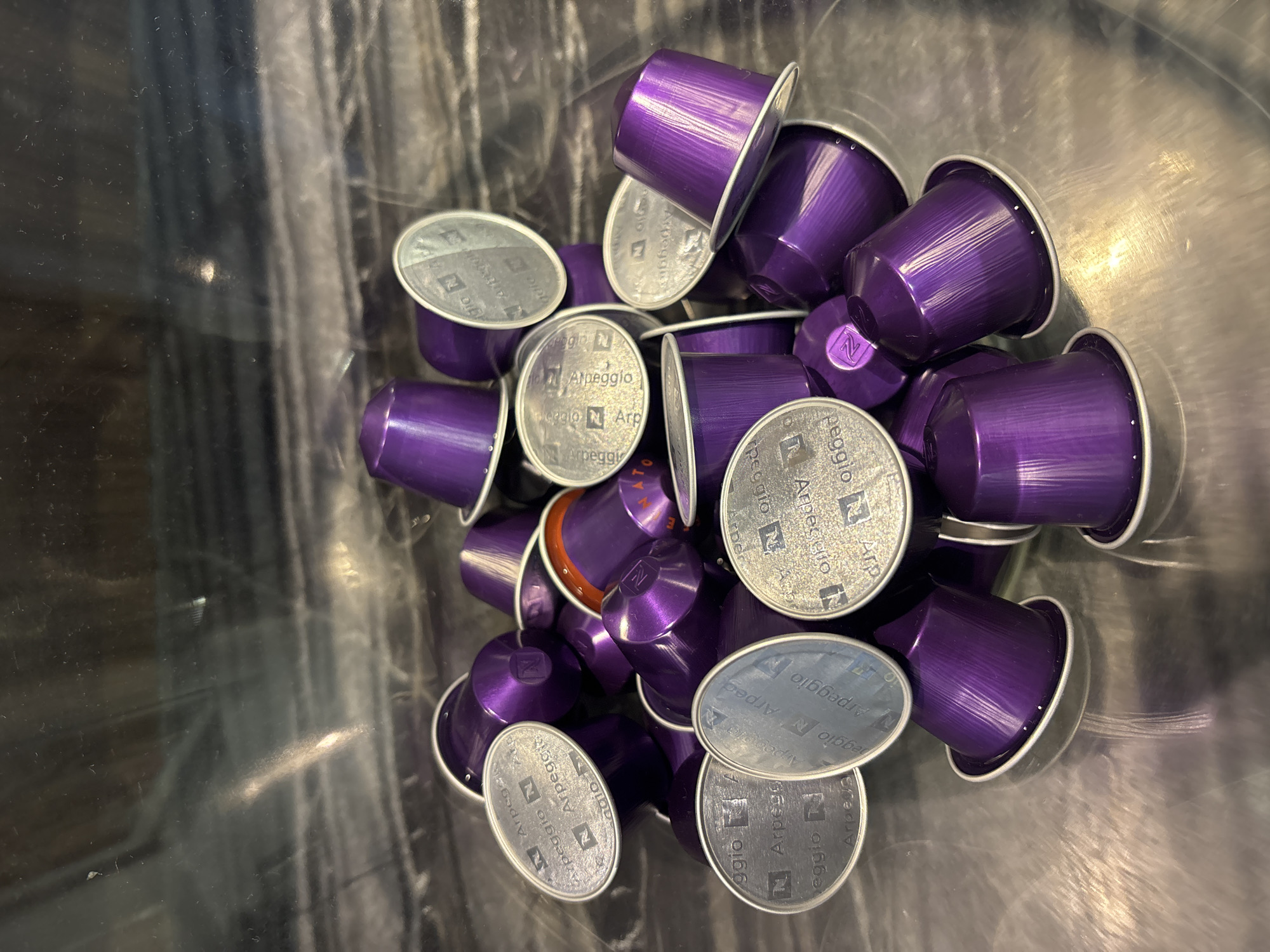 A close-up overhead shot shows a pile of Nespresso Arpeggio coffee capsules. The capsules are predominantly purple with silver foil lids that feature the Nespresso logo and "Arpeggio" text. Some of the capsules are partially obscured by others. The lighting is somewhat harsh, creating reflections on the metallic surfaces of the capsules and the clear container they are in. The background is out of focus, showing a dark, textured surface that suggests a countertop or shelf. There are no people or any indication of a specific location within Dubai, United Arab Emirates.