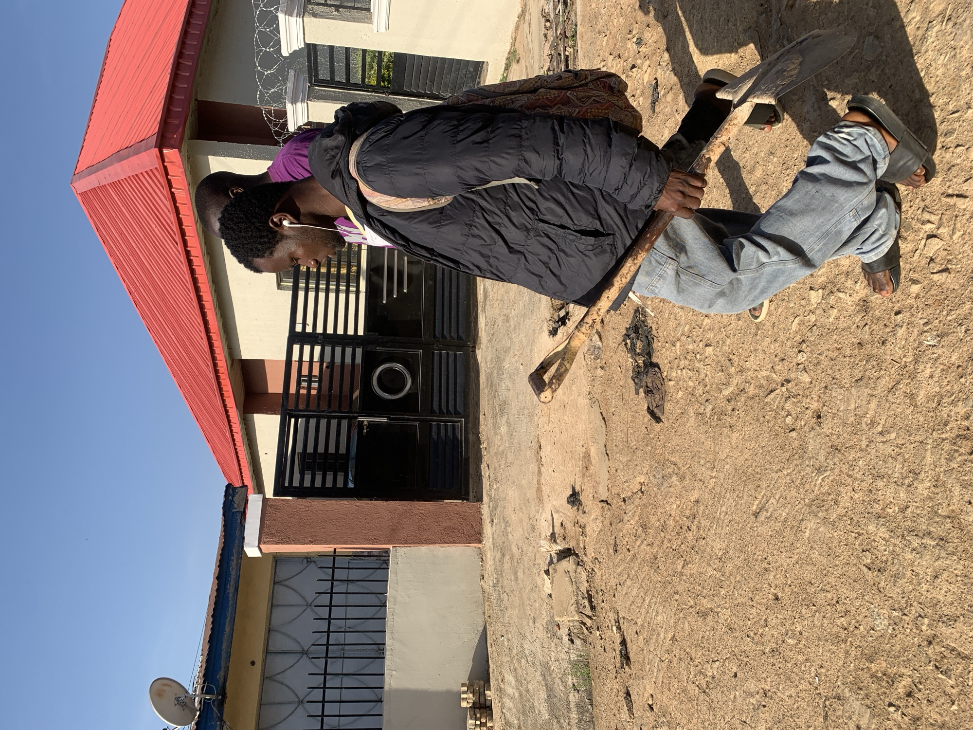 A man stands in front of a building on a dirt road in Gura Top, Nigeria. He is carrying a shovel on his shoulder and wearing blue jeans, a black jacket, and sandals. He has headphones in his ears. The building has a red roof and a brown and tan facade. There is a satellite dish on the side of the building.