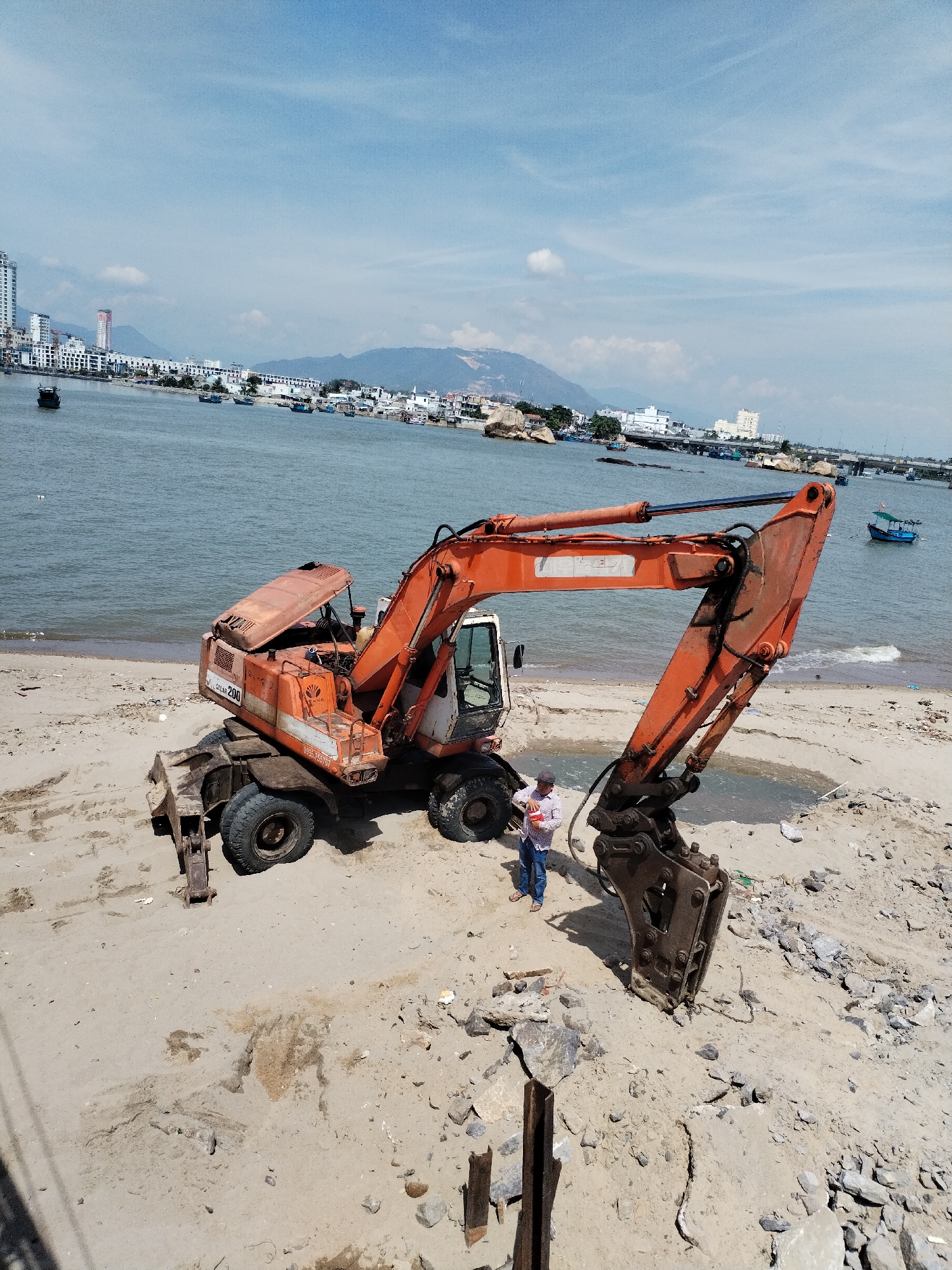 An orange excavator is parked on a sandy beach next to the sea in Bac Nha Trang Ward, Vietnam. The excavator's engine cover is open, revealing some mechanical components. A person in a white shirt and blue jeans stands near the excavator, holding a red and white object. The sea is calm, with several boats visible in the distance. The background shows a cityscape with buildings and mountains. The sand is dry and uneven, with some debris scattered around. The sky is partly cloudy.