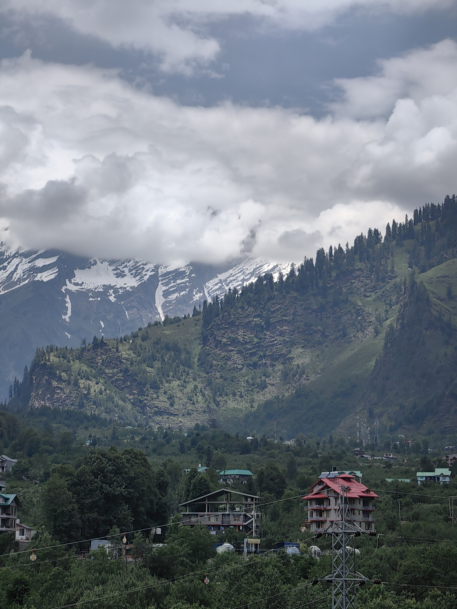 The image depicts a mountainous landscape with snow-capped peaks under a cloudy sky. In the foreground, there are several houses nestled among lush green trees. The houses vary in size and architectural style, with one prominent building featuring a red roof and multiple stories. A utility tower stands near the red-roofed house, with power lines stretching across the scene. The overall environment appears to be a rural or semi-urban area situated in a hilly region, likely Kulang, India, given the context. The scene is static, with no visible people or actions. The lighting is subdued due to the cloud cover.