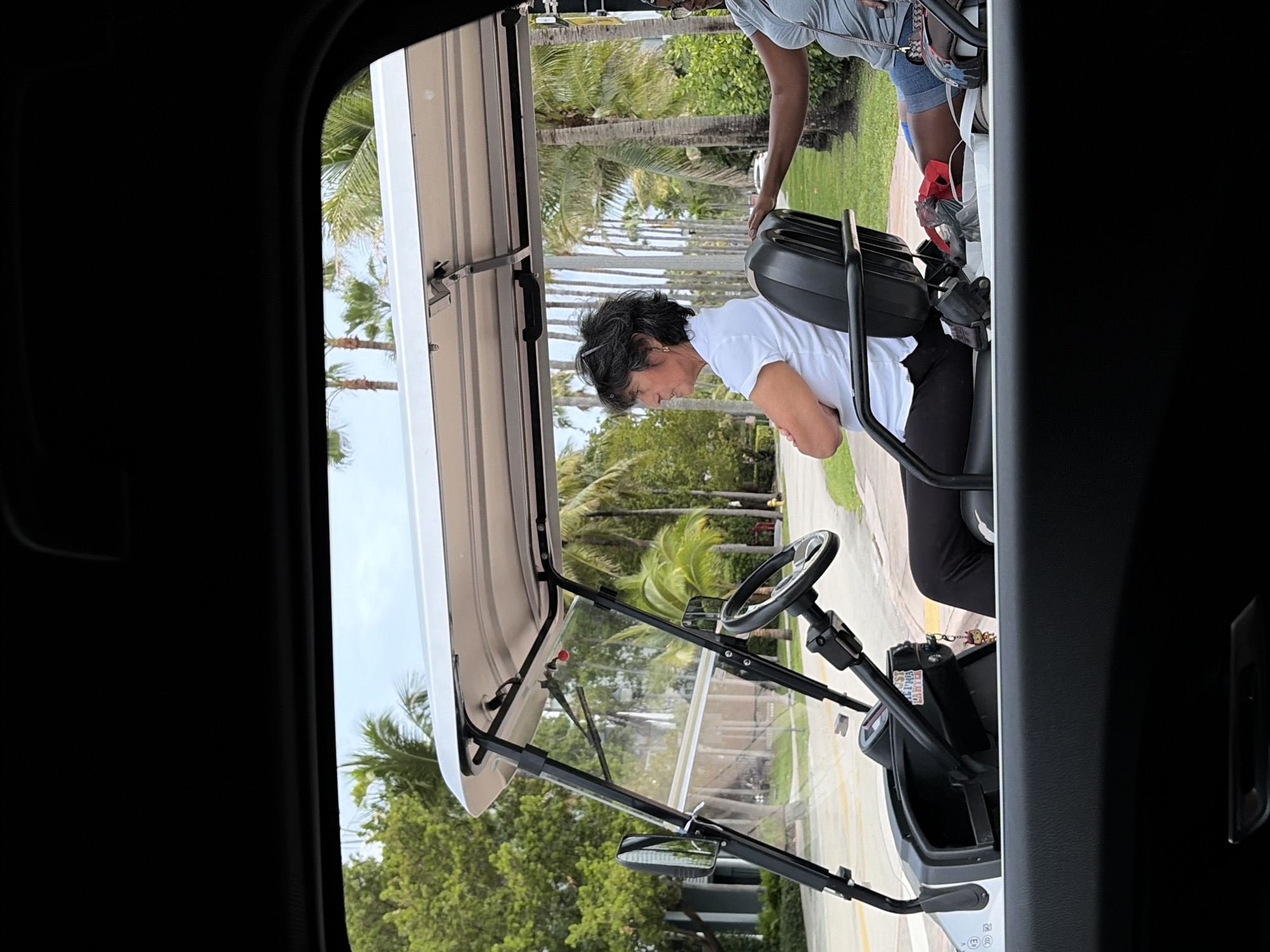 The image, taken from within a vehicle, shows a woman sitting in a golf cart. She wears a white shirt and dark pants and has dark, short hair. Her arms are crossed. Another person is partially visible standing near the golf cart, wearing a gray shirt and denim shorts. The golf cart is white with a black steering wheel and frame. In the background, there is a row of palm trees lining a street. The image appears to have been taken in the Key Biscayne area, United States of America, evidenced by the prevalence of palm trees.
