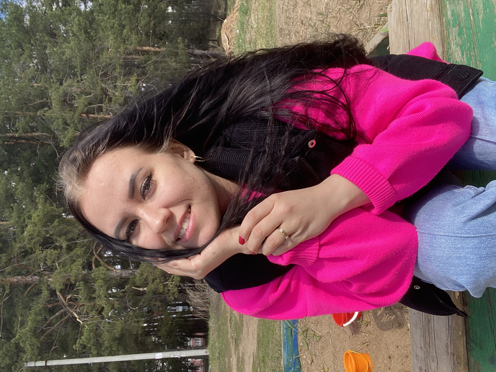 A young woman is seated outdoors, possibly on a wooden structure with painted green areas. She is wearing a bright pink sweater over a black jacket, with blue jeans. She has long, dark hair with lighter streaks, and is smiling with her hands near her face. She has red nail polish on the visible fingernails and wears a gold ring on one finger. The background shows trees and a patch of ground. Orange and white objects, possibly children's buckets, are on the ground near her feet. Based on the environment (trees) it is likely a city park, possibly located in the city of Chaikovsky, Russia.