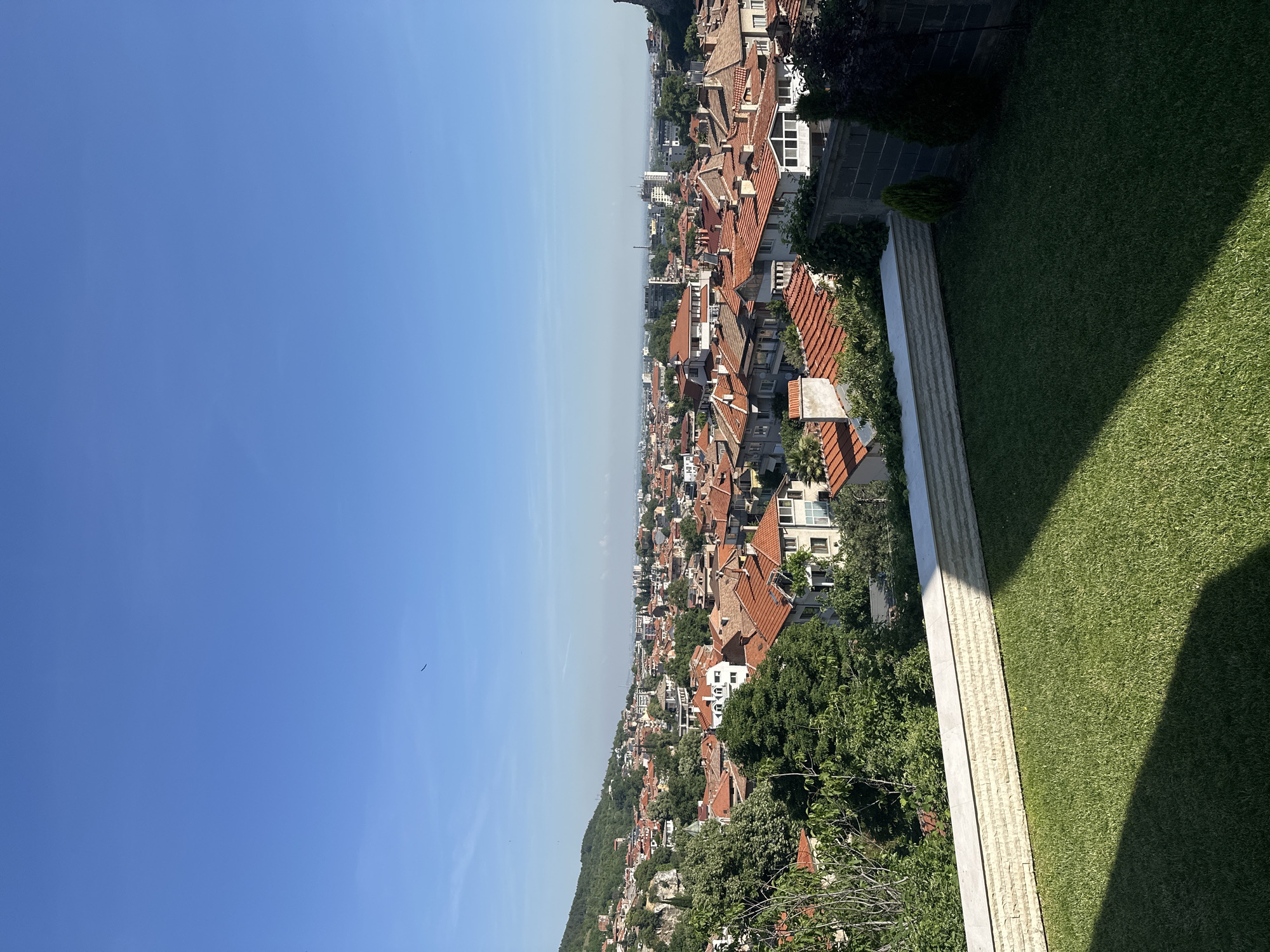The image captures a panoramic view of Asenovgrad, Bulgaria, showcasing a dense urban landscape characterized by terracotta-tiled rooftops. The foreground features a neatly manicured green lawn with a shadow cast from an unseen object, suggesting it is a sunny day. A low, decorative wall made of light-colored stone separates the lawn from the backdrop of the city.
The midground is dominated by a cluster of buildings with predominantly red-tiled roofs, indicative of traditional architecture. Some buildings exhibit white facades, while others are a mix of muted colors. Lush green trees are interspersed throughout the city, adding a natural element to the urban sprawl.
In the background, the cityscape extends towards the horizon, with taller, more modern buildings visible in the distance. A hazy, light blue sky stretches across the top of the frame, with a few faint clouds and what appears to be a bird in flight. To the left, a green, tree-covered hill slopes upwards, with some buildings nestled into its side. To the far right, a rocky outcrop or hillside rises, partially obscuring the city beyond. The overall impression is that of a vibrant city on a clear day, viewed from an elevated vantage point.
