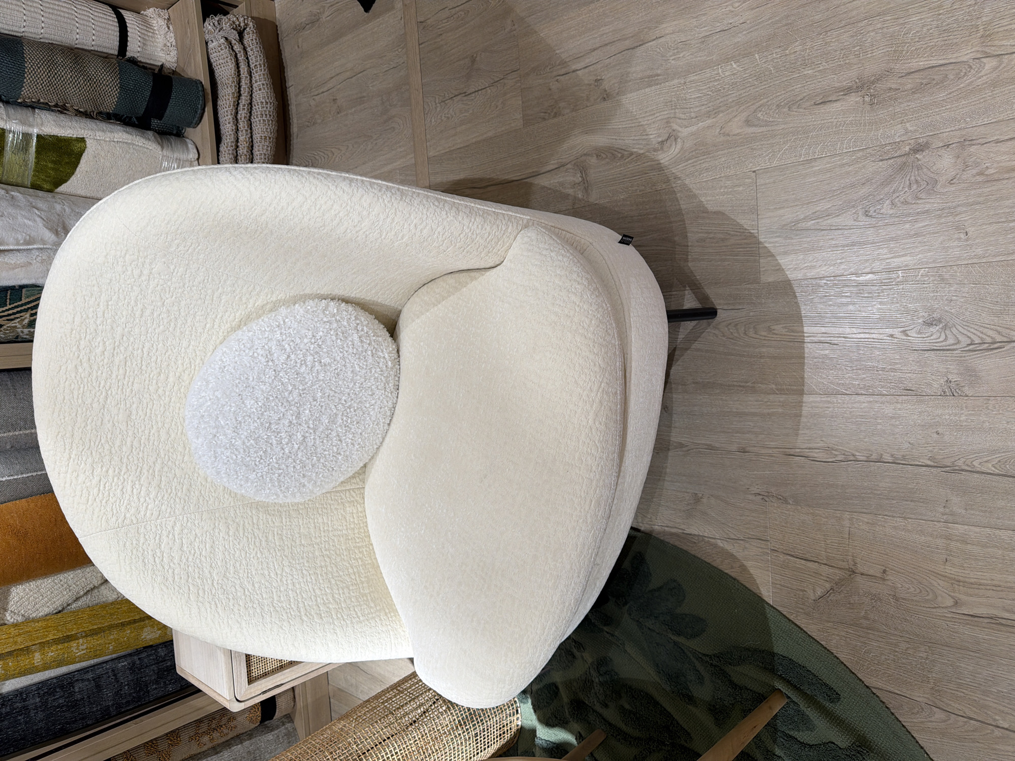 The image shows a plush, cream-colored armchair with a round, textured white pillow on its seat. The armchair has a curved back and seat, and rests on thin black legs. In the background, there are several rolled-up rugs and folded textiles displayed on shelves. To the left of the armchair, a woven basket and a dark green rug with a raised pattern are partially visible. The floor is made of light brown wooden planks. The scene appears to be an interior setting, possibly a retail store or showroom. There are no people or animals present.