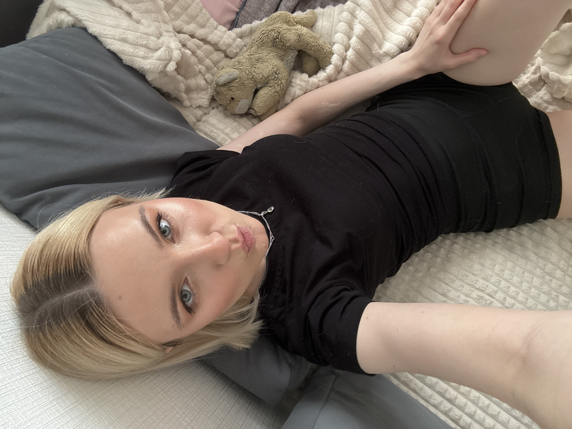 A woman lies on a bed with a grey pillow and a beige blanket. She wears a black t-shirt and dark shorts. A stuffed animal is on the blanket beside her. The image is a close-up selfie taken from above. There is no information to determine a location in Vsevolozhsk, Russia.