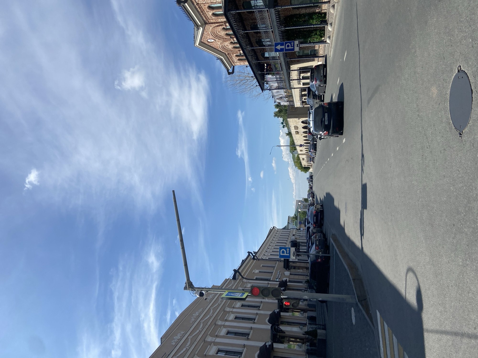 The image depicts a wide-angle view of a sunny urban street in Kazan, Russia. The street extends into the distance, flanked by multi-story buildings on both sides under a bright blue sky with scattered white clouds.
On the left side of the street, a light beige building with ornate cornices and decorative window frames lines the sidewalk. Several dark awnings are visible over ground-floor entrances. A gray utility pole stands in the foreground, supporting a horizontal arm extending over the street, to which a CCTV camera is attached. Below the camera, a red traffic light for vehicles and a red pedestrian signal (standing figure) are visible. A blue pedestrian crossing sign (white walking figure) is also affixed to the pole. A marked pedestrian crosswalk with white zebra stripes is visible on the asphalt in front of the pole. Several dark-colored cars are parked along the curb on this side of the street. Further down, a blue square parking sign with a white "P" and symbols indicating paid parking is present.
On the right side of the street, a reddish-brown brick building features numerous arched windows and elaborate black wrought-iron balconies on its upper floors. Another blue parking sign with an upward-pointing arrow is visible near this building. Further down the street on the right, a long, lower building with a stone-colored facade and a series of arched openings is visible. Above this structure, green domes, consistent with religious architecture, are discernible in the distance. Several cars are also parked along the curb on this side.
The asphalt road surface features dashed white lane markings. A black car with a partially visible Russian license plate ("048AEA 16") is in the foreground, moving away from the viewer. Other cars are visible both parked and in motion further down the street. A dark, circular metal manhole cover is present on the road surface in the immediate foreground. Long shadows from the buildings and street furniture stretch across the road, indicating a high sun angle. No people are discernible in the scene.