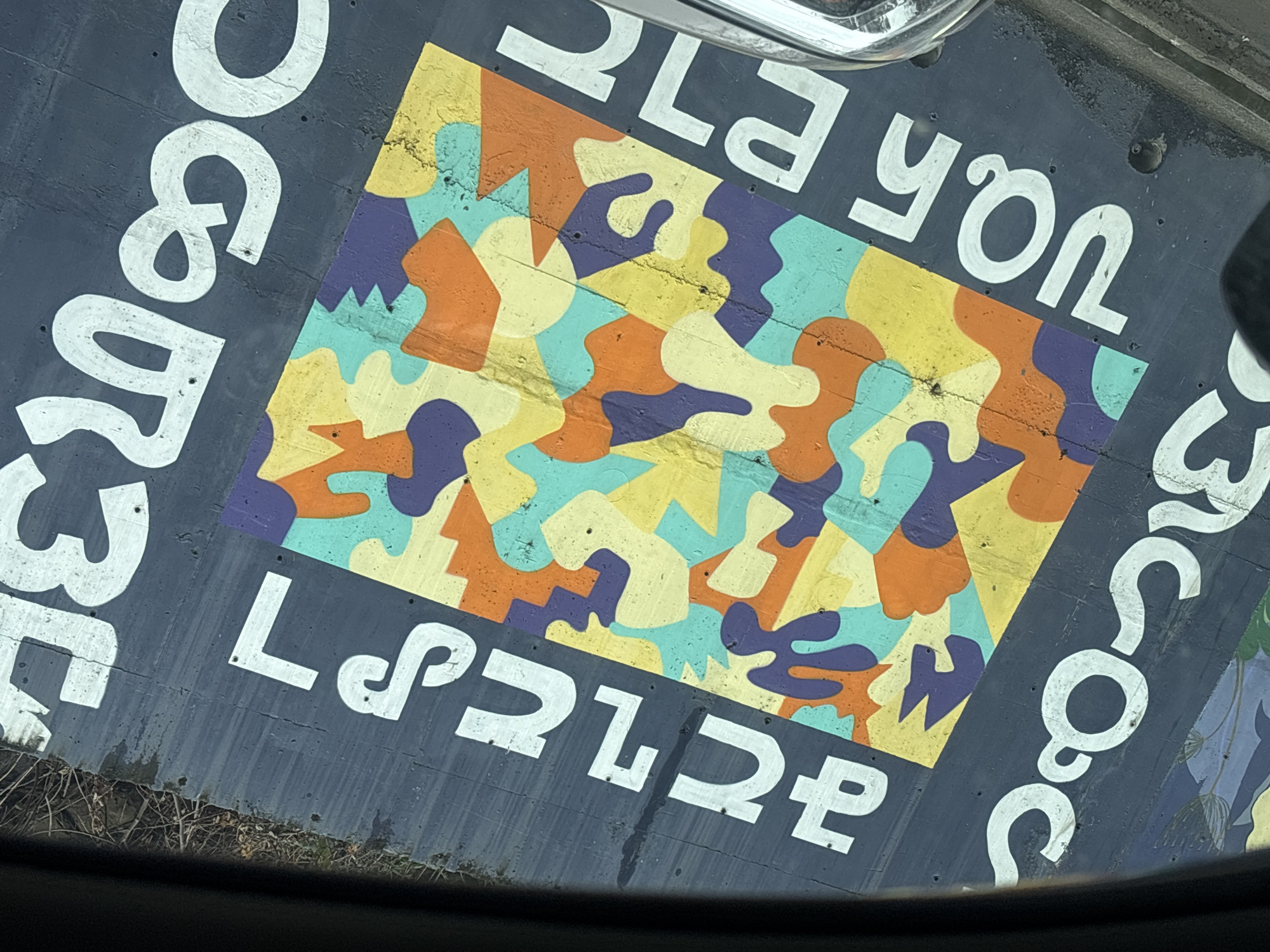 The image displays a section of a concrete mural painted on a wall along Tamar Mepe Avenue, specifically at the underpass near Didube Metro Station in Tbilisi, Georgia. The photograph is taken from inside a vehicle, evidenced by the dark, curved frame of a windshield or dashboard partially obscuring the top and bottom foreground.
The mural features a central rectangular panel filled with an abstract, tessellating pattern composed of irregular, rounded shapes in yellow, light blue/teal, orange, and dark purple. This central design is bordered by a dark blue-grey concrete surface. White Georgian script (Mkhedruli) is painted on this surrounding surface, spelling out four words: "სიყვარულს" (Siyvaruls - Love) in the top left quadrant, "მშვიდობას" (Mshvidobas - Peace) in the top right, "თავისუფლებას" (Tavisuplebas - Freedom) in the bottom left, and "მეგობრობას" (Megobrobas - Friendship) in the bottom right, framing the abstract artwork.
The concrete surface exhibits visible texture, minor irregularities, and what appear to be small drill holes and subtle streaking or staining. Above the upper edge of the wall, a patch of dry, sparse vegetation is partially visible. No individuals or discernible actions are depicted in the static scene.