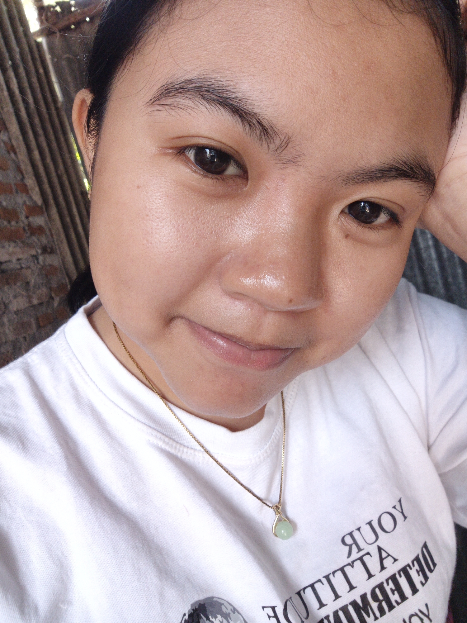 A young woman with dark hair and brown eyes is shown from the chest up, looking directly at the camera. She is wearing a white t-shirt with a gold chain and a light green pendant necklace. The background is blurry, but appears to be an outdoor setting with a wooden fence and brick wall. The image is a close-up selfie, with the woman's right hand resting on the side of her head.