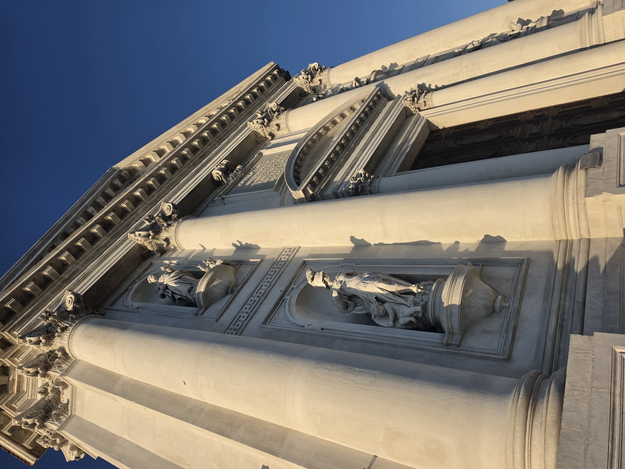The image shows a low-angle view of the facade of a grand white building, likely a church or public building, with ornate classical architecture. The building features large fluted columns, intricate carvings on the entablature, and niches containing white marble statues. One statue depicts a female figure in armor with a helmet, accompanied by a smaller cherubic figure. Another statue, higher up, shows a robed female figure. The building is bathed in the warm light of the sun, casting strong shadows on its surfaces. The sky above is a clear, vibrant blue. The architectural style suggests it is located in Venice, Italy, possibly the Church of Santa Maria della Salute due to its prominent dome, columns, and statues, though the facade does not fully reveal the dome. The scene is static, with no people visible.