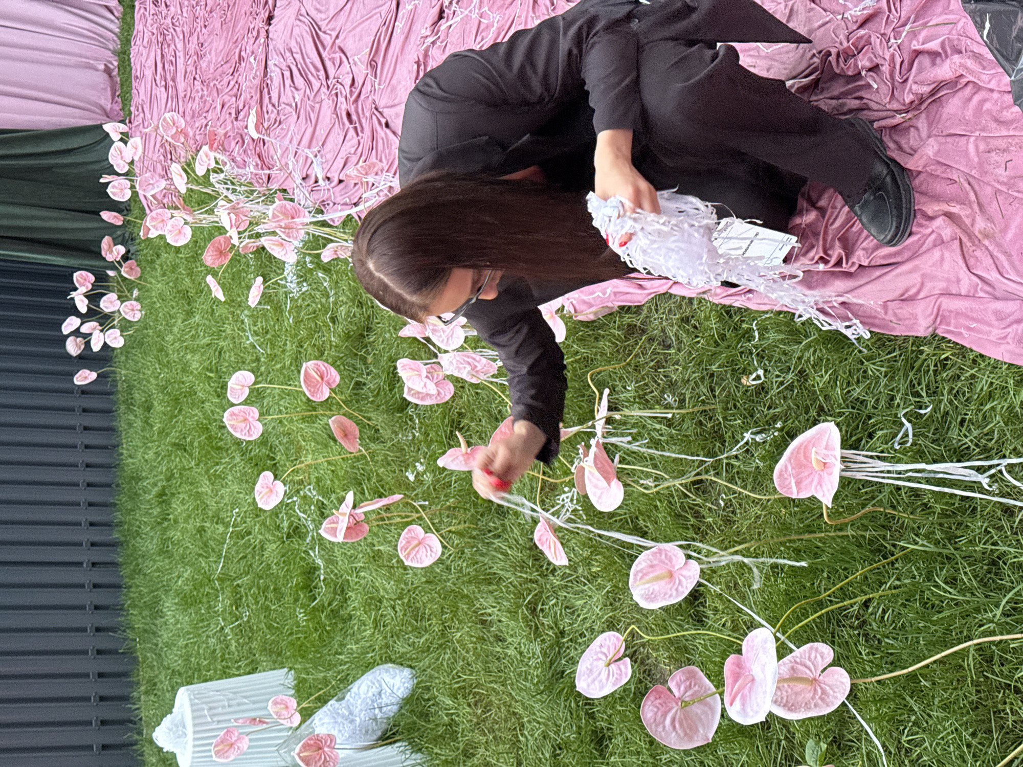 In the city of Perm, Russia, a person dressed in black is crouched on a pink fabric, tending to pink calla lilies arranged on a grassy area. The person, with long dark hair and glasses, holds a cluster of white shredded paper in their hands. Several pink calla lilies are scattered across the lawn, some with white streamers attached to their stems. In the background, a dark grey fence borders the grassy area, with green and pink draped fabric formations visible further back. To the left, a white fluted object and another object wrapped in white material are also present on the grass.