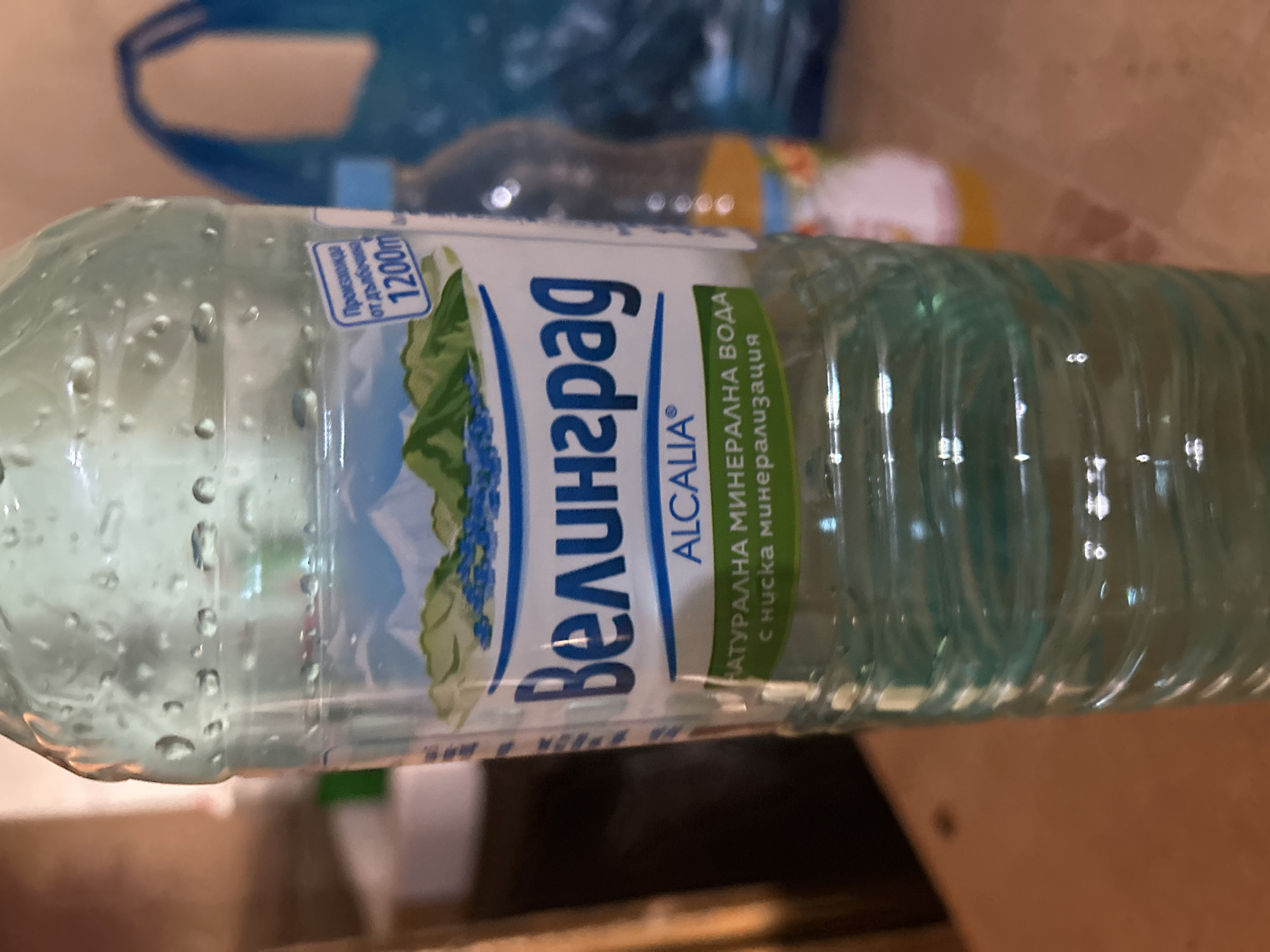 A clear plastic bottle of mineral water, likely cold due to the visible condensation and water droplets on its surface, is depicted lying horizontally. The background is out of focus, showing indistinct shapes and colors, including muted blue, yellow, and brown tones, suggesting an indoor setting with other objects.
The bottle features a prominent label with the brand name "ВЕЛИНГРАД" (VELINGRAD) in large blue Cyrillic letters. Below the main brand, the text "ALCALIA®" is visible. To the right of the brand name, green text indicates "НАТУРАЛНА МИНЕРАЛНА ВОДА с ниска минерализация" (NATURAL MINERAL WATER with low mineralization). On the upper left portion of the label, a blue-outlined, cloud-like graphic states "Произхожда от дълбочина 1200m" (Originates from a depth of 1200m). The label also incorporates a visual design of green mountainous terrain with blue flowers. The bottle itself has a textured surface with vertical ridges. The water inside appears clear and still.