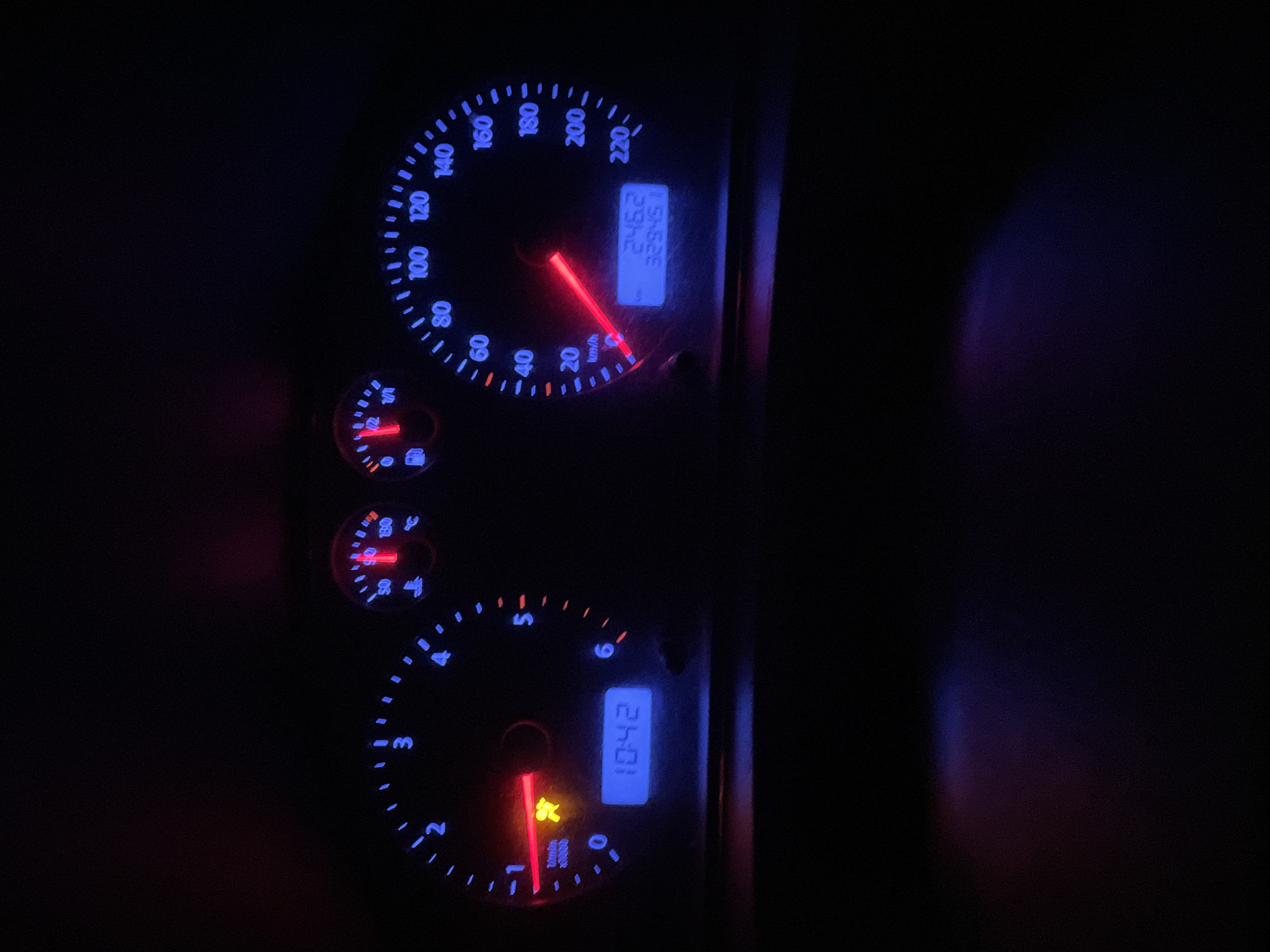 The image captures a car's instrument cluster at night, illuminated with a characteristic blue backlighting and red needles against a dark background. The dashboard features several gauges and digital displays.
The speedometer, located on the upper right, indicates speeds up to 220 km/h. Its needle points to 0 km/h, suggesting the vehicle is stationary or moving very slowly. A digital display next to the speedometer shows a trip odometer reading of "245.2" km and a total odometer reading of "329451" km. Below these, a fuel gauge indicates the tank is almost empty, accompanied by an illuminated red low fuel warning light. A smaller gauge for engine temperature is also visible, currently showing a low reading.
On the lower left, the tachometer displays engine revolutions per minute, ranging from 0 to 6 x1000 /min. Next to the tachometer, a second digital display shows the time as "10:42". A prominent red warning light, resembling an airbag or seatbelt reminder symbol, is illuminated below the tachometer needle.
The scene is set inside a car in Moscow, Russia, during nighttime, evidenced by the darkness surrounding the brightly lit dashboard. No individuals are visible. The car's ignition appears to be on, as indicated by the active displays and warning lights.