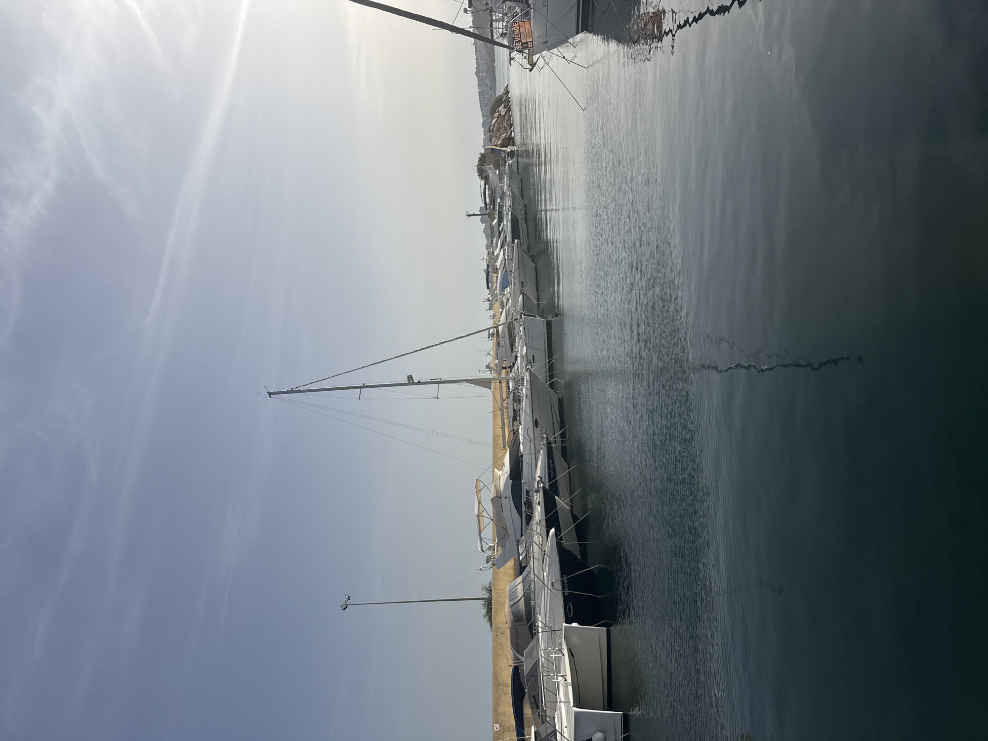 The image shows a marina filled with various yachts and sailboats docked along a pier. The water is calm and reflects the sky and the boats. The sky is clear with a few wispy clouds and the sun is shining brightly, casting a gentle glare on the water. In the background, there is a rocky outcrop with some buildings and vegetation on top, suggesting a coastal city or town. A tall lamppost is visible near the pier, and some of the boats have covers on them. One boat in the foreground on the right has a "SE VENDE" sign on it, indicating it is for sale.