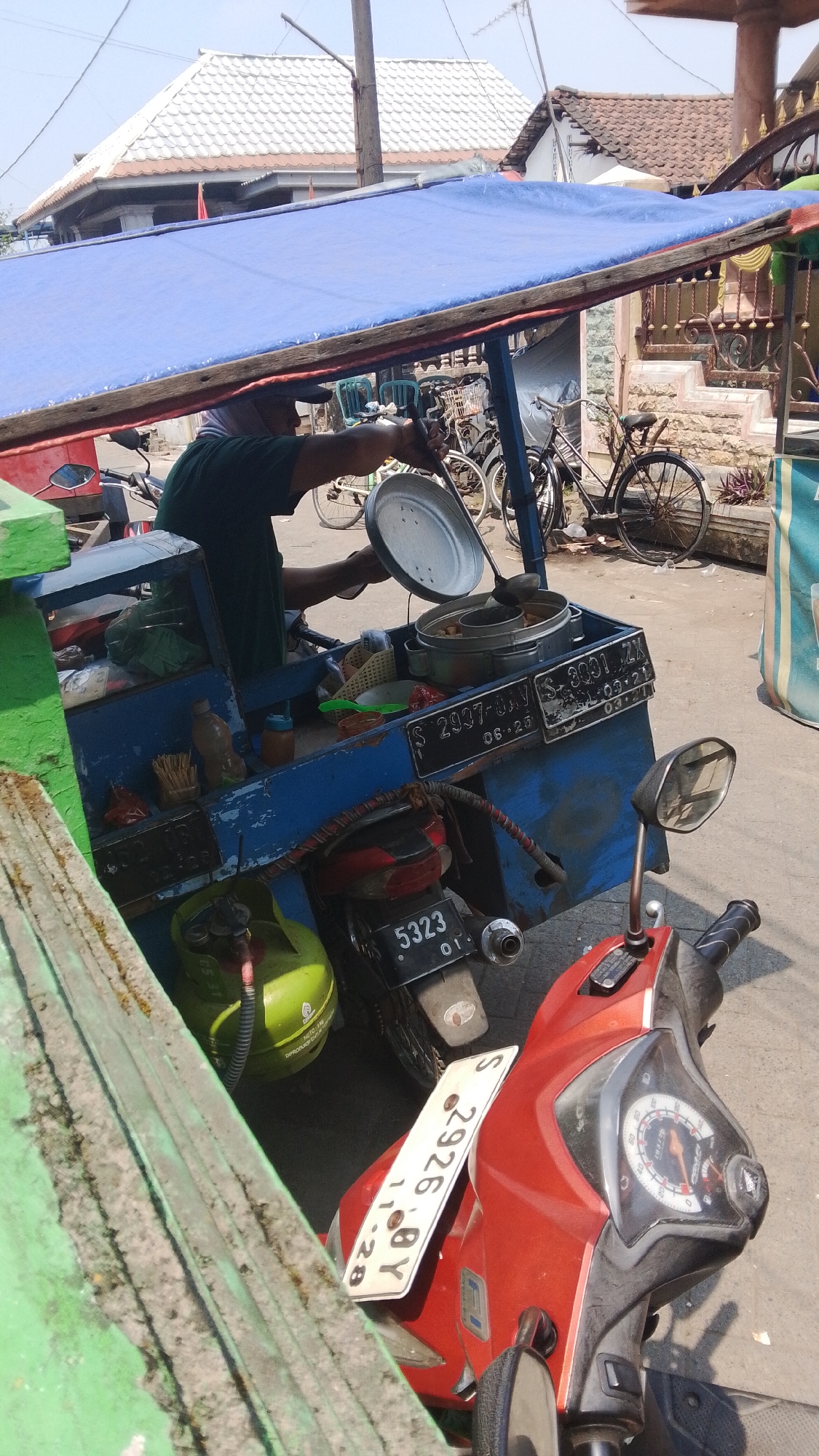In Mancar, Indonesia, a street vendor is preparing food from a mobile cart, likely on a sunny day. The cart, painted blue, is mounted on a vehicle or trailer and covered with a large blue tarp. A person, wearing a green shirt and a head covering, is actively serving food from a metal pot using a ladle. Several metal pots and containers are visible on the cart, suggesting a food preparation area. A green propane tank is attached to the cart, indicating a cooking source. Behind the cart, various bicycles are parked. In the foreground, a red motorcycle is parked, with a white license plate visible. The background shows buildings with tiled roofs and a street. The overall scene depicts a typical street food vending operation in an Indonesian setting.