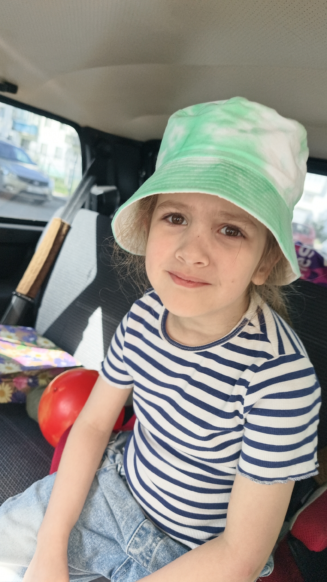 A young child with fair skin, light brown hair, and brown eyes is seated inside a car. The child wears a white and blue striped shirt, light blue denim shorts, and a light green and white tie-dye bucket hat. To the child's left, behind the seat, is a tan wooden stick with a black joint piece resting on top of a light gray seat cover, alongside a red ball and a patterned bag. The car's interior is visible, and through the windows, a blurred urban scene is visible, suggesting the location may be Chaikovsky, Russia. The child is looking towards the camera with a slight smile.