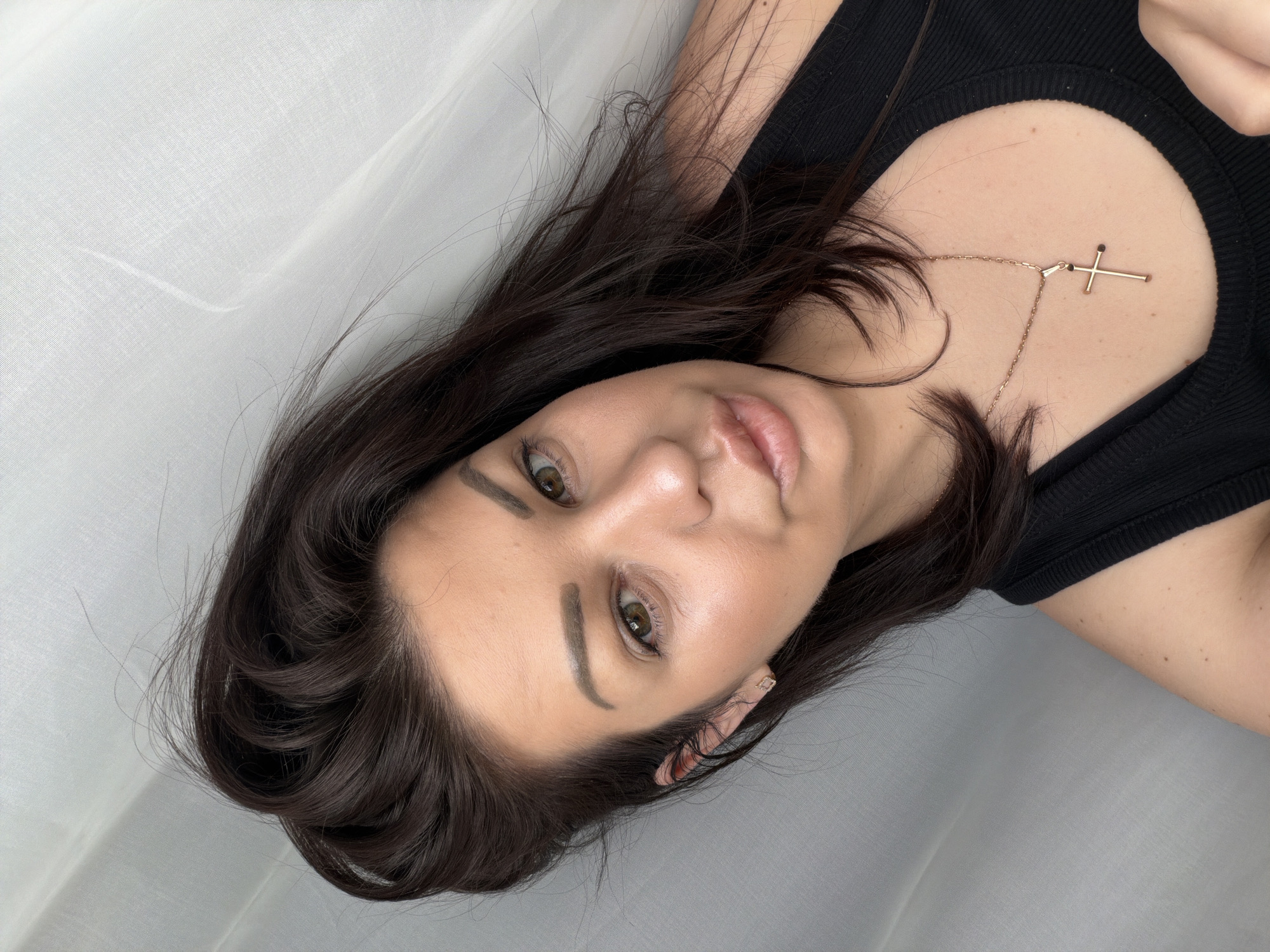This is a close-up, waist-up, selfie-style photograph of a woman with dark brown hair and light brown eyes. She is wearing a black ribbed tank top and a gold cross necklace. Her hair is styled loosely, with some strands falling across her face and shoulders. She is looking towards the camera with a neutral expression, and her lips are slightly parted. The background is a softly draped white fabric, suggesting an indoor setting. The lighting appears to be natural, coming from the left, creating a gentle glow on her face. There is no indication of the city of Sochi, Russia, or any specific location within it.