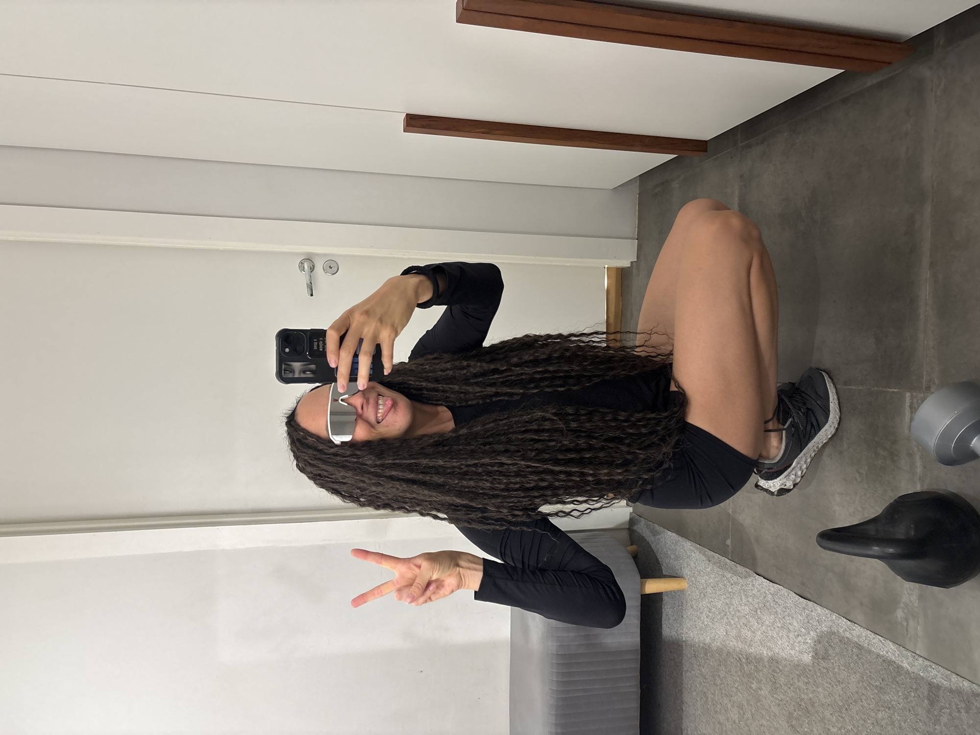 A person with long, dark, textured hair, wearing silver-framed sunglasses, a black long-sleeved top, and black shorts, takes a mirror selfie while squatting or crouching indoors. They are smiling broadly and holding a smartphone, which features a design of eyes on its case, while making a peace sign with their left hand. They are wearing black athletic shoes with white soles.
The setting appears to be a modern indoor space in Chaikovsky, Russia, possibly a home gym or fitness area, as indicated by the presence of a grey dumbbell and a black kettlebell on the floor. The background visible in the mirror reflection includes a white wall or door with a visible door handle, and white ceiling or wall panels accented by two horizontal wooden beams. A grey, concrete-like wall or floor is also visible. A grey upholstered bench with wooden legs is partially seen in the reflection. The overall atmosphere is casual and cheerful. Text on the phone case is partially visible, appearing to include words like "STARK" and "POWER."