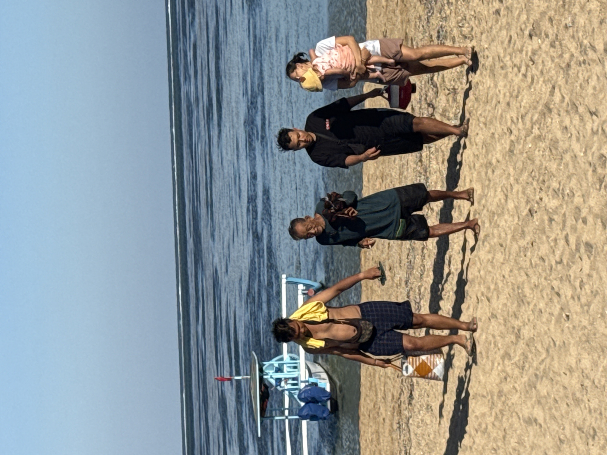 A group of four people walks along a sandy beach in Denpasar, Indonesia. The ocean is calm with gentle waves and a clear sky above. In the background, a traditional Balinese fishing boat with a blue and white structure is moored in the shallow water.
On the left, a man with a yellow towel draped over his shoulders walks, carrying a patterned bag and flip-flops. He is wearing checkered shorts. Beside him, an older man in a dark teal shirt walks, holding what appears to be a small object in his hands.
In the center, a man in a black t-shirt and black shorts walks with a red cooler bag. To his right, a woman carries a young child, who is wearing a yellow sun hat and a pink outfit. The child is nestled in the woman's arms, facing away from the camera. The woman wears a white t-shirt and brown shorts. The group is moving from left to right across the frame. The sand is light brown and shows some footprints. The lighting suggests it is daytime, likely morning or afternoon, with clear shadows cast on the sand.