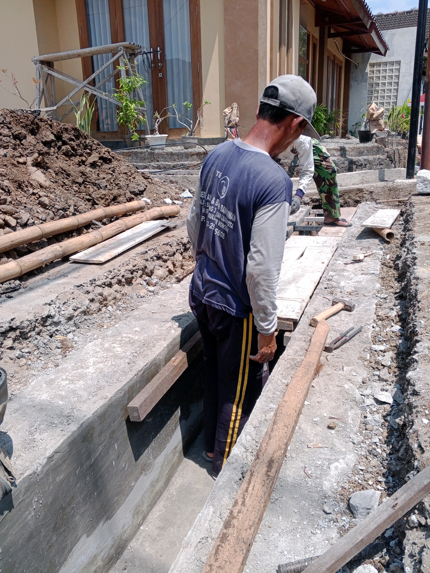 The image depicts a construction site in Giritirto, Indonesia, with two male laborers present. One laborer, wearing a grey cap and a dark blue long-sleeved shirt with a logo, stands facing away from the camera, observing the work. The other laborer, in camouflage pants and a light-colored shirt, is partially visible in the background, also engaged in the construction. The scene shows an excavated trench, with concrete lining and wooden planks laid across it for access. Soil, bamboo poles, and construction debris are scattered around the site. The environment suggests an ongoing infrastructure project, possibly related to drainage or utility installation.