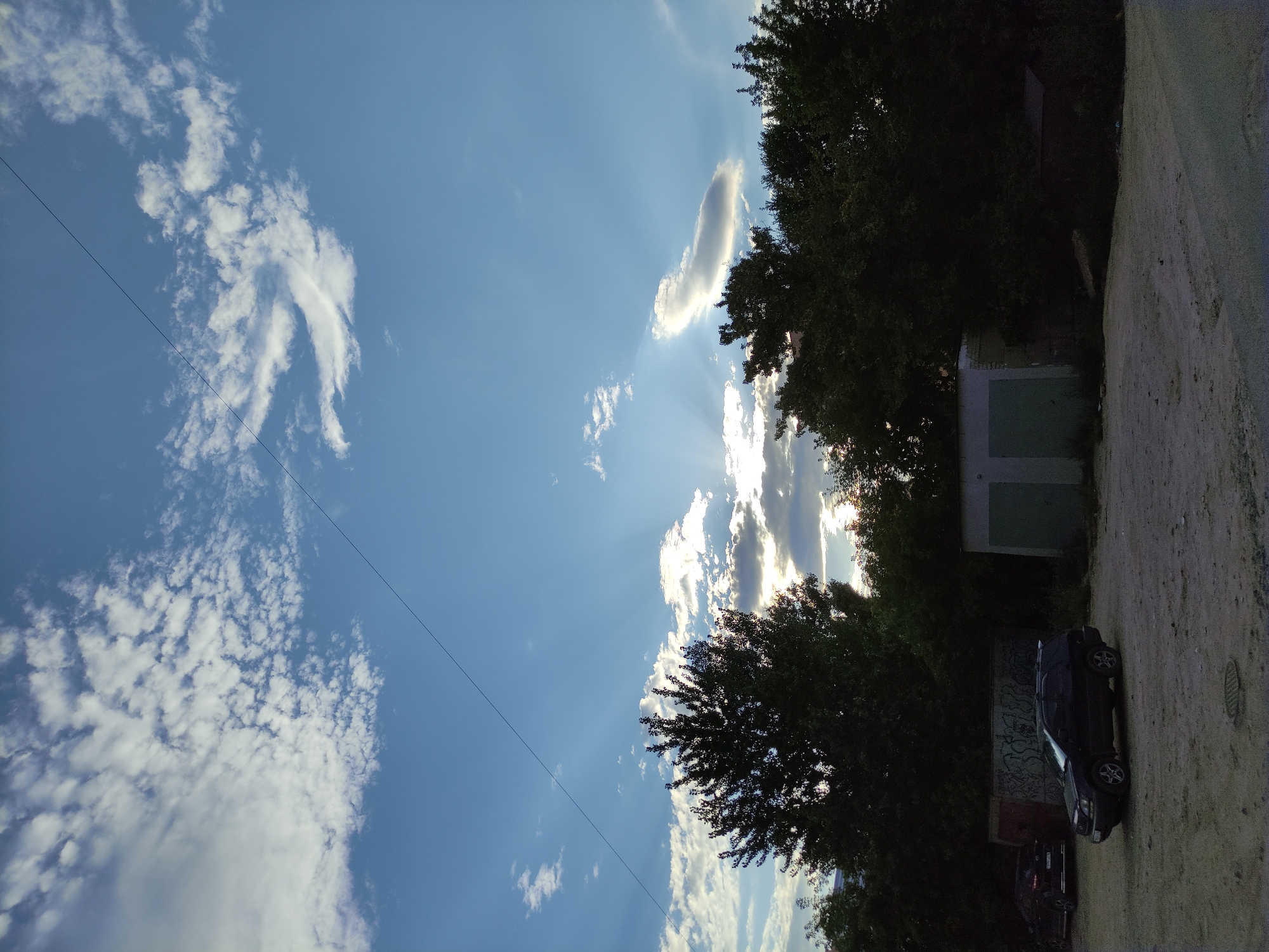 The image captures a sky with scattered clouds and visible sun rays emanating from behind them, suggesting it is either sunrise or sunset. The sun appears to be low on the horizon, casting light in a diffused manner. Silhouetted against the sky are the tops of several trees, their leaves dense and dark. In the foreground, a dirt or gravel area is visible, with two dark-colored cars parked. One car, a dark SUV, is in the center of the frame, while another is parked further back on the left. Behind the cars and partially obscured by trees, there are several garages or sheds with light-colored doors. A thin, dark power line stretches diagonally across the upper portion of the sky. The overall scene depicts an outdoor urban or semi-urban environment, likely a parking area adjacent to vegetation and small structures, under a dramatic sky.