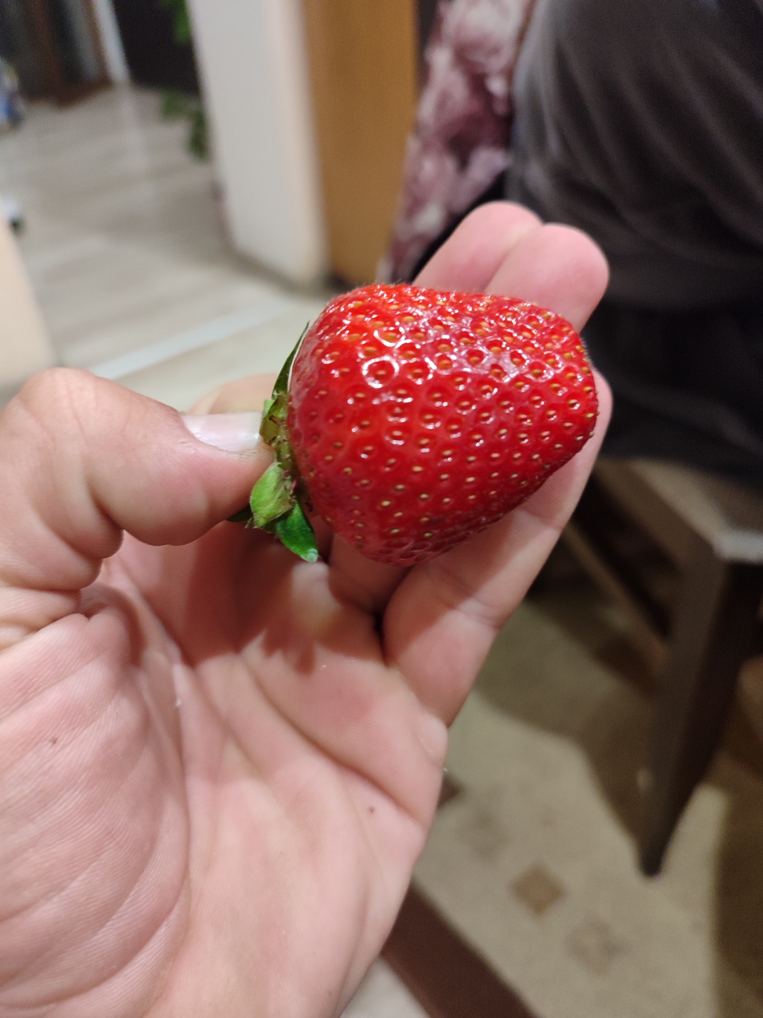 A fair-skinned human hand holds a bright red strawberry with green leaves at the top. The strawberry is ripe and covered in small seeds. The hand appears slightly wrinkled. The background is blurred but shows a floor, a portion of a dark chair, and a wall. The location is undetermined.
