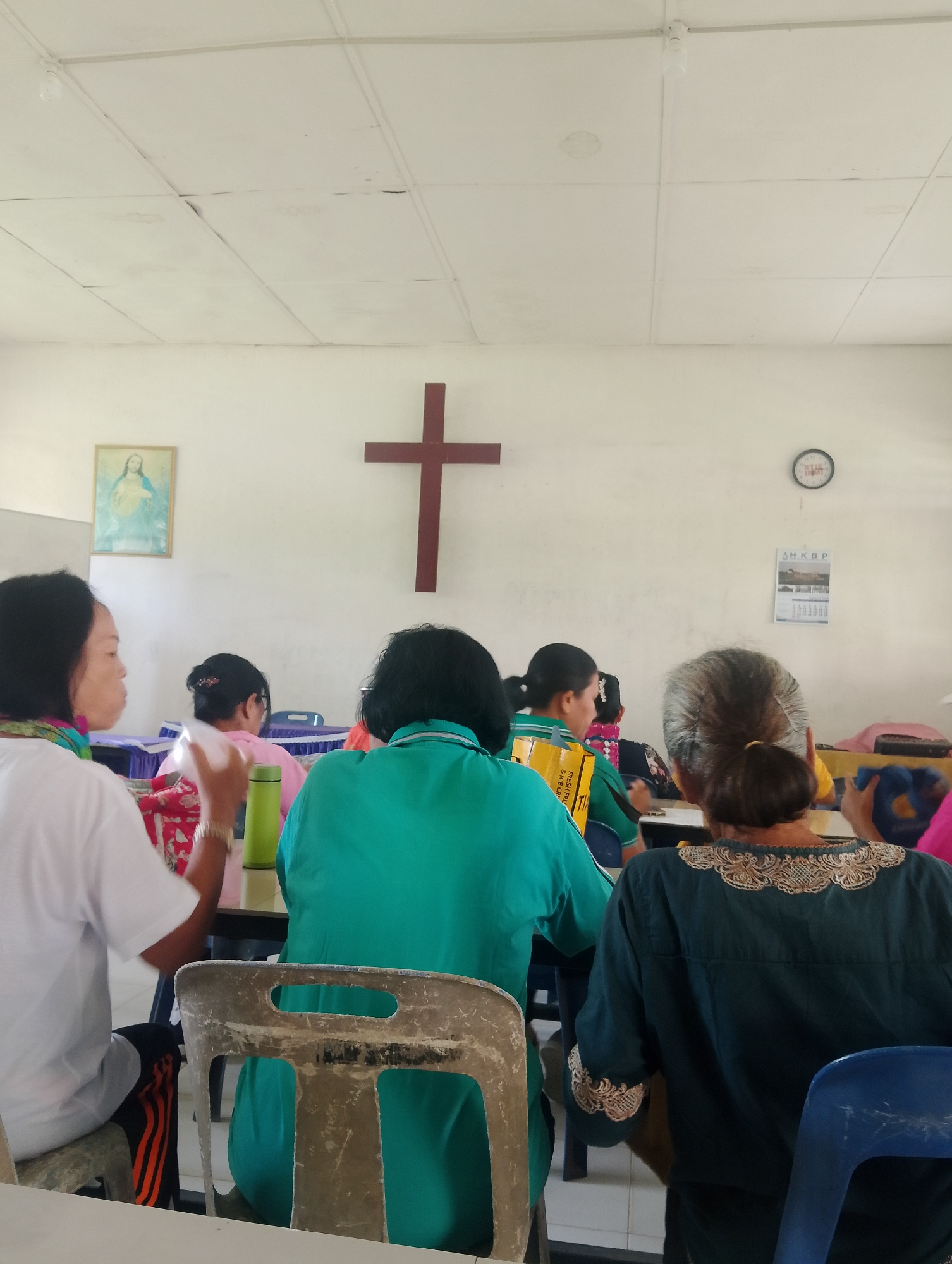 In Serbelawan, Indonesia, a group of women are seated in what appears to be a community hall or meeting room. The room is simply furnished with white walls and a white drop ceiling. A large wooden cross is prominently displayed on the back wall, suggesting a religious affiliation, possibly a church or a Christian community center. To the left of the cross, a framed image of Jesus Christ is also visible on the wall.
Several women are seated at tables, facing away from the camera. One woman in the foreground on the left wears a white t-shirt and has a colorful scarf around her neck. Another woman in the center wears a green t-shirt and appears to be holding a green water bottle. A woman further back is wearing a pink top. The woman in the immediate foreground on the right is wearing a dark blue long-sleeved shirt with decorative embroidery on the collar and sleeves. The environment suggests a gathering for a religious service, bible study, or a community event hosted by a church. The lighting appears to be natural and from overhead fixtures.