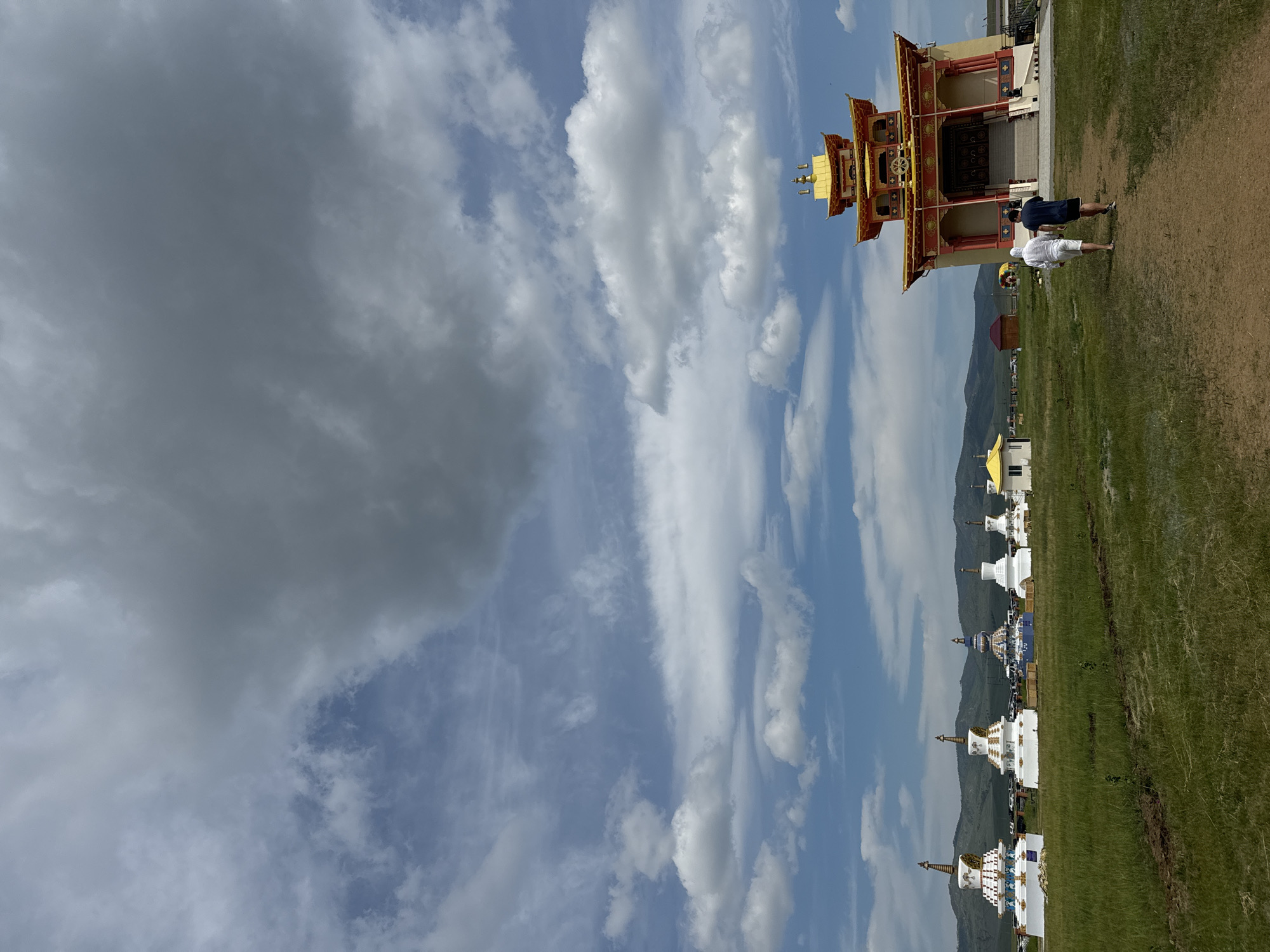 In Orongoi, Russia, a sunny day with scattered clouds frames a Buddhist complex. A golden-roofed, red and yellow temple stands on the right, its entrance accessed by a set of stairs. Two men, one in a white hooded sweatshirt and shorts, the other in a dark t-shirt and shorts, walk away from the camera towards the temple. In the middle ground, a line of white stupas, some adorned with gold and blue accents, stretches across a grassy field. A smaller white building with a yellow roof is visible to the left of the temple, and distant mountains form the backdrop under a dynamic sky.