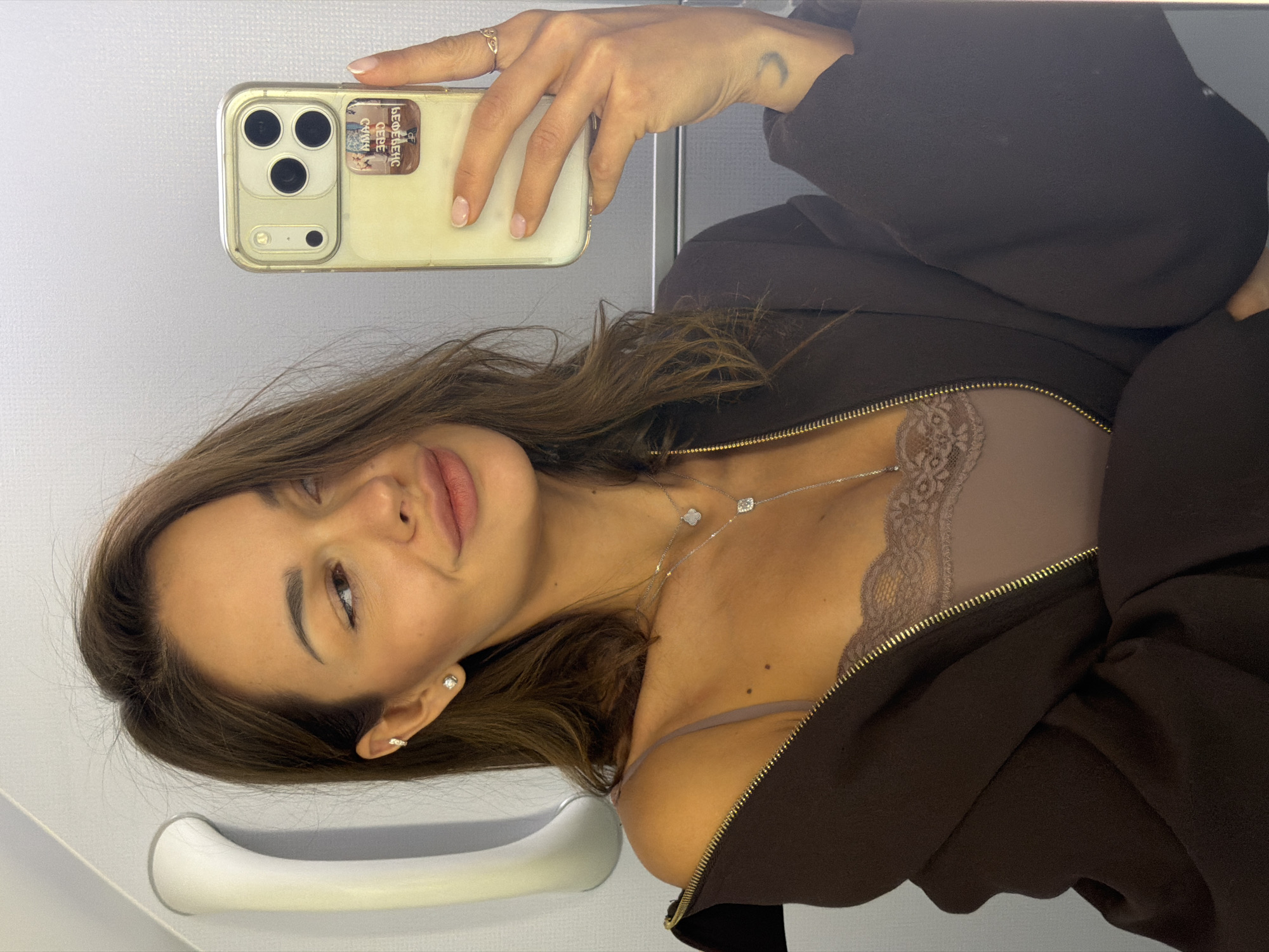 A woman is taking a mirror selfie in what appears to be the interior of an airplane. She has medium-length brown hair, and is wearing a brown open jacket over a taupe camisole with lace trim. She has on delicate silver necklaces, and two small diamond stud earrings, one in her lobe and one higher on her ear. On her left wrist is a small crescent moon tattoo. She is holding a smartphone, the screen displaying a sticker of what appears to be a vintage advertisement. The background shows a plain, light-colored wall and a white handrail.