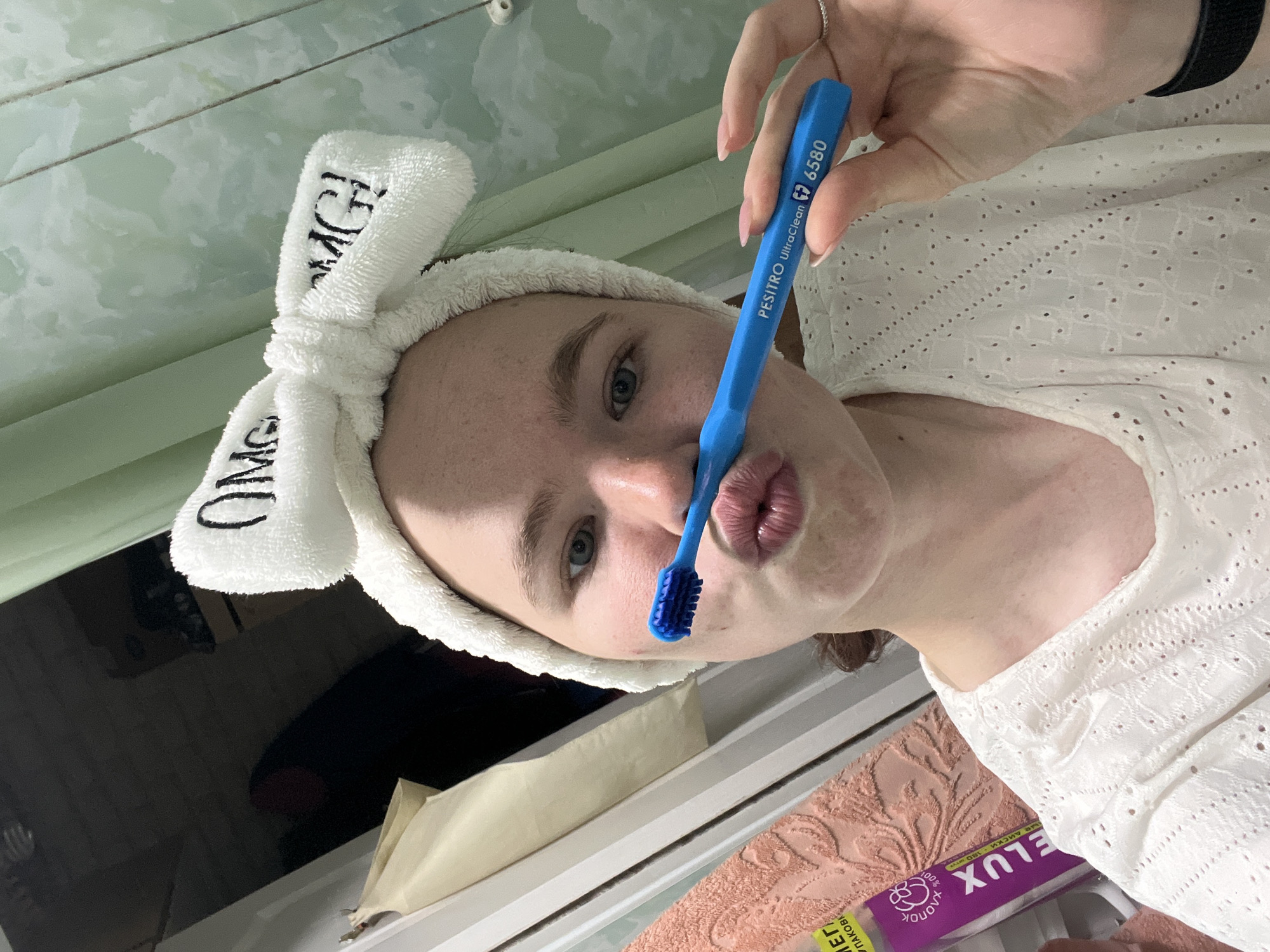 A person with fair skin is posing with a blue toothbrush. The person is wearing a white headband with the word "OMG" written on it, and a white eyelet top. The toothbrush is blue and has the words "PESITRO UltraClean 6580" printed on it. The background appears to be a bathroom, with green-tiled walls and a dark, possibly black object that looks like a TV or mirror. A beige box and what appears to be a pink patterned towel are also visible. A product in purple packaging with the word "DELUX" in reverse is in the bottom right corner. The photo was taken in Chaikovsky, Russia.
