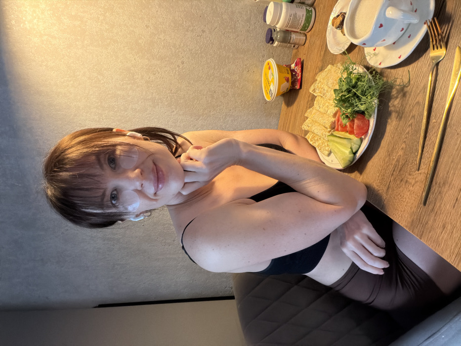 The image displays a woman seated at a wooden table indoors, located in Perm, Russia. She faces the viewer, smiling faintly. The woman has medium brown hair with bangs and is wearing two circular, transparent under-eye patches. White wireless earbuds are visible in both ears, and she wears small hoop earrings. Her left hand rests under her chin. She is dressed in a black spaghetti strap crop top and dark brown fitted shorts. She is seated in a dark brown upholstered chair.
On the table, a white plate holds several triangular pieces of flatbread, sliced green cucumbers, sliced red tomatoes, and a mound of green microgreens. To the right of the plate, a white cup with a red heart-patterned saucer contains a light brown beverage. A gold-colored fork and knife are positioned on the table in front of the plate. Behind the main plate, a yellow plastic container is visible next to a small red foil-wrapped item. Multiple plastic bottles, likely containing supplements or medications, and a small dark spray bottle are also present. A smaller white plate partially visible holds a dark item. The background consists of a textured, light gray wall, illuminated by a warm light source on the right side.