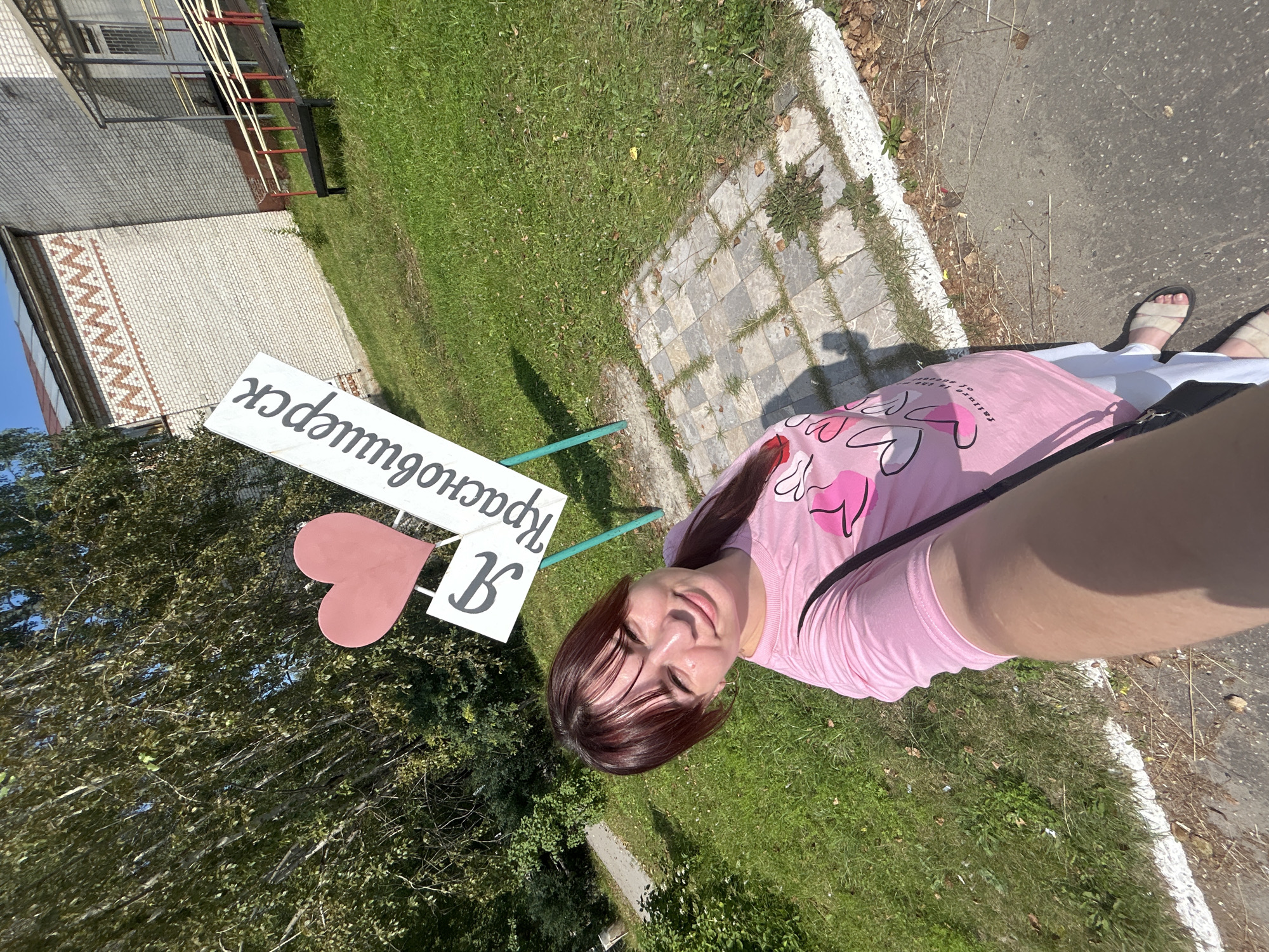 In an outdoor setting, a young woman with brown hair and a pink t-shirt is taking a selfie. She is standing in front of a sign that reads "Я люблю Красновишерск" (I love Krasnovishersk) with a heart symbol. The sign is white with black lettering and is mounted on a green pole. Behind the sign, a brick building with a decorative band of zig-zag pattern is visible, along with a metal staircase leading up to an entrance. The ground is covered with grass and a paved path made of square tiles. Trees are present on the left side of the image. The weather appears to be sunny, with a clear blue sky. The overall scene suggests a location within the city of Krasnovishersk, Russia, likely a public space or park.