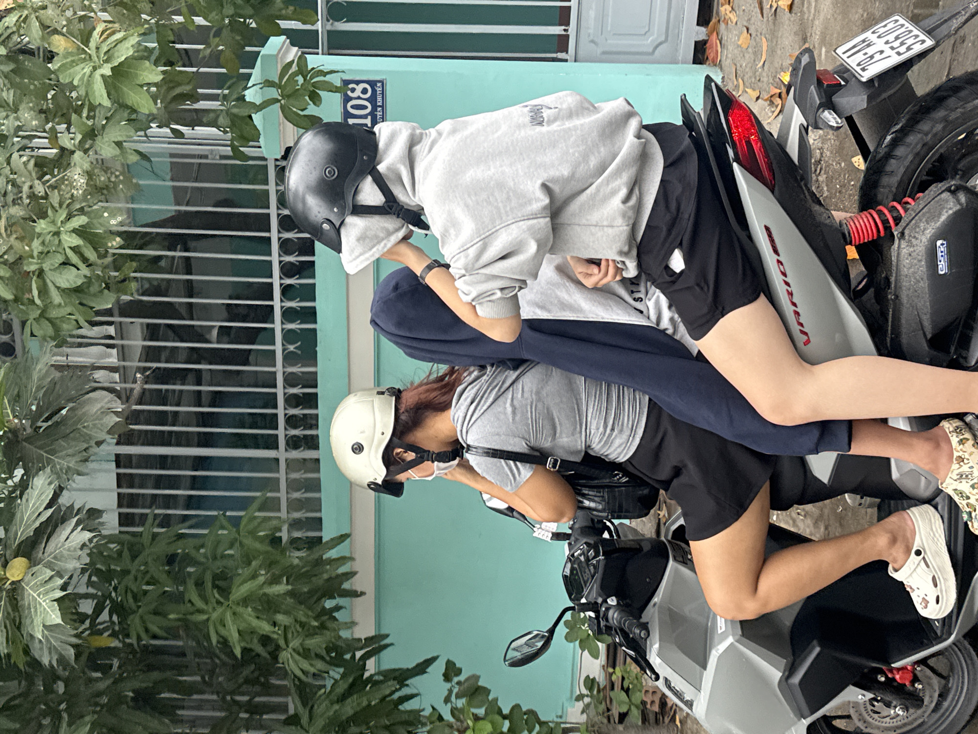 Two individuals are riding on a silver motorcycle in Nha Trang, Vietnam. The person in the front is wearing a white helmet, a gray t-shirt, and black shorts. They have a black backpack and are wearing a white mask. The person in the back is wearing a black helmet and a grey hooded sweatshirt with black shorts. They are holding a dark blue garment over their body. The motorcycle is on a paved road with fallen leaves scattered around. In the background, there is a teal-colored building with a white metal fence and some greenery. A house number "108" is visible on the building. The license plate of the motorcycle is "79-AA 556.03".