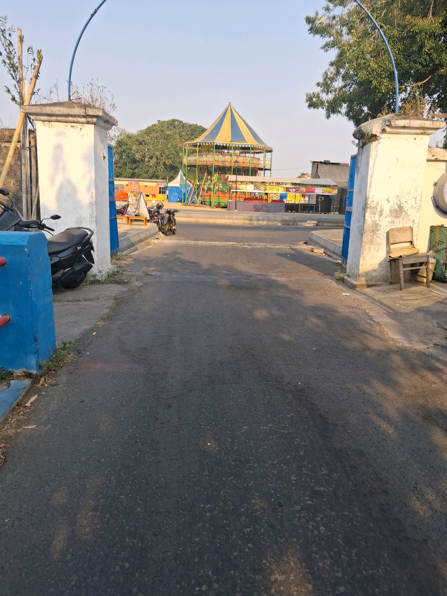 The image shows an outdoor scene in Surakarta, Indonesia, likely near an amusement area. The perspective is from a slightly elevated position looking forward through an archway or entryway supported by two large white pillars. The pillars have weathered surfaces and are part of a structure that includes blue metal gates.
In the foreground, a paved asphalt road leads into the scene. To the left, a blue concrete barrier is visible. A black motorcycle is parked to the left of the road, partially obscured by a pillar. On the right side of the road, near the pillar, a rustic wooden chair with a light-colored cushion sits on the ground.
Beyond the entryway, the scene opens up to a bustling area. A multi-level amusement ride with a distinctive yellow and blue striped conical roof is prominent in the background, featuring colorful structures and decorations. Several market stalls or temporary structures with awnings and signs are set up in front of the ride. A few more motorcycles are parked in this area. Trees and other vegetation are visible in the middle ground and background, with a clear sky overhead, suggesting it is daytime. The sunlight casts shadows across the road, indicating the sun is at an angle.