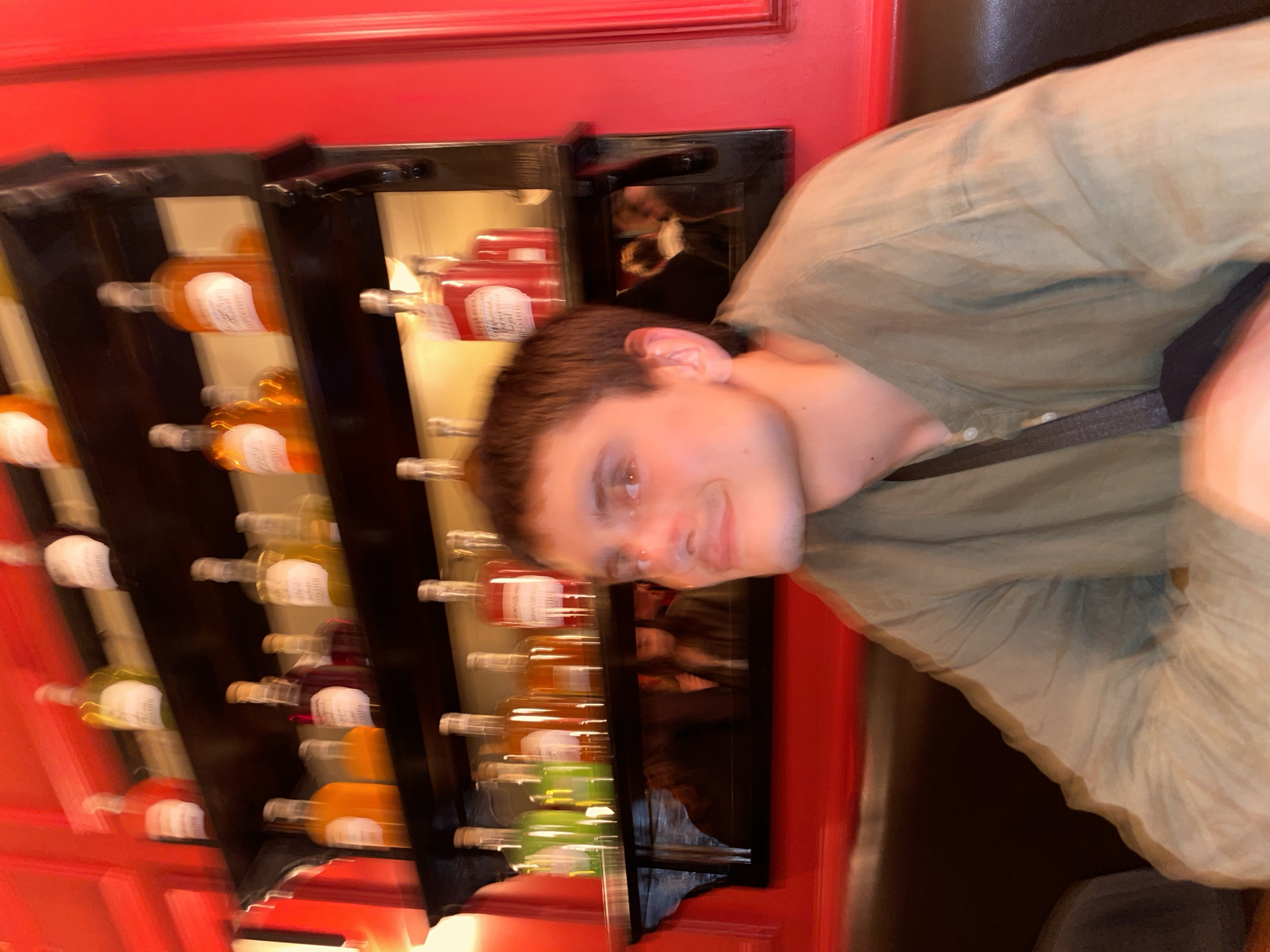 A slightly blurred image shows a fair-skinned young man in a green shirt standing next to a wine rack against a red wall. The wine rack holds numerous bottles of wine, many with white labels. Another person's reflection is visible in the rack's glass panel. The setting is possibly Moscow, Russia, considering the context of the request.