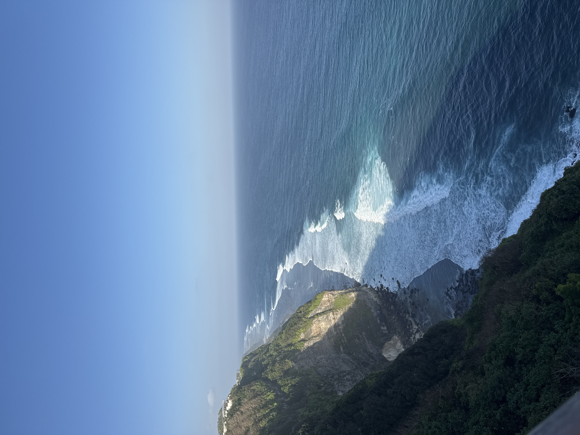 A vertical, high-angle, wide shot captures a breathtaking coastal cliff overlooking a vast ocean. The sky above is a clear, gradient blue, transitioning from a lighter hue near the horizon to a deeper shade at the top. Sunlight casts a dramatic diagonal shadow across the cliff face, highlighting its rugged texture and verdant green vegetation. The cliff plunges steeply into the sea, where powerful white waves crash against the rocky shore. Beyond the breaking waves, the ocean stretches out, a mesmerizing expanse of deep blue water that gradually lightens to turquoise closer to the shore. In the upper left corner of the frame, a cluster of white buildings is visible perched atop the cliff, hinting at human presence in this otherwise wild landscape. The scene evokes a sense of raw, natural beauty and immense scale.