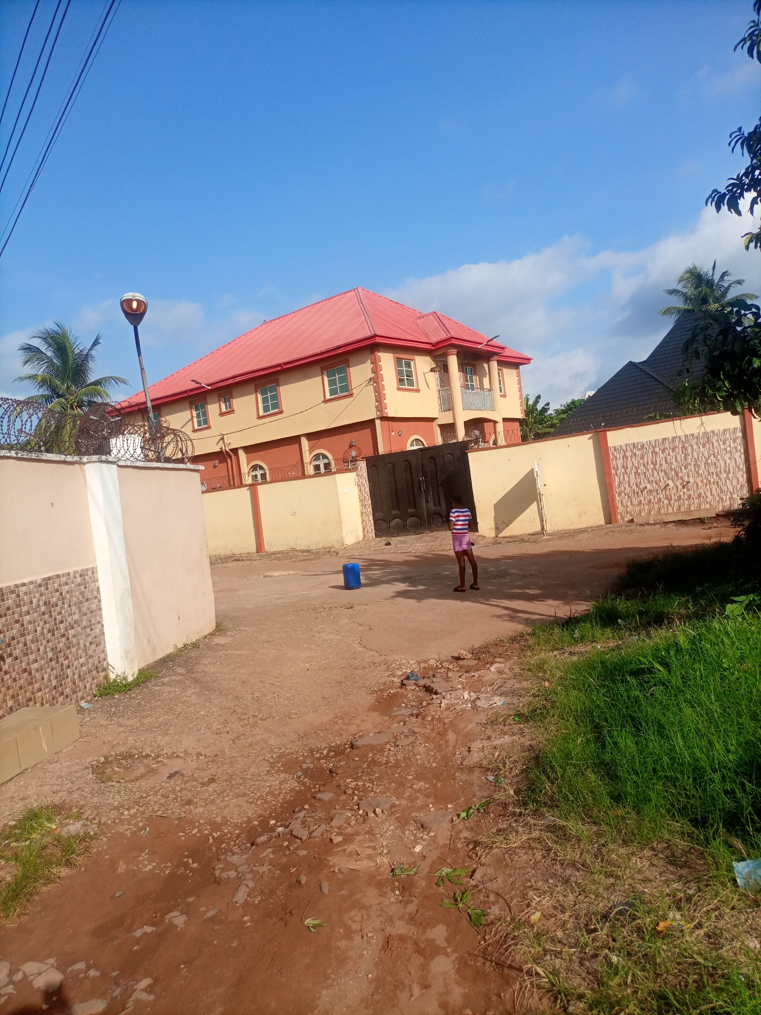 An exterior, low-angle, eye-level shot captures a two-story building with a red roof in Nkpor, Nigeria, under a bright blue sky with scattered clouds. The building has a cream-colored facade with reddish-brown accents and multiple windows with white frames. A large black gate stands in front of the building. A person wearing a striped t-shirt and shorts stands on a dirt path in front of the gate, facing away from the camera. The path is uneven and shows signs of wear and tear. A tall street lamp is visible to the left of the building, and barbed wire tops a light-colored wall in the foreground. Green grass and foliage border the path on the right side of the image. A blue plastic container is situated on the path near the building.