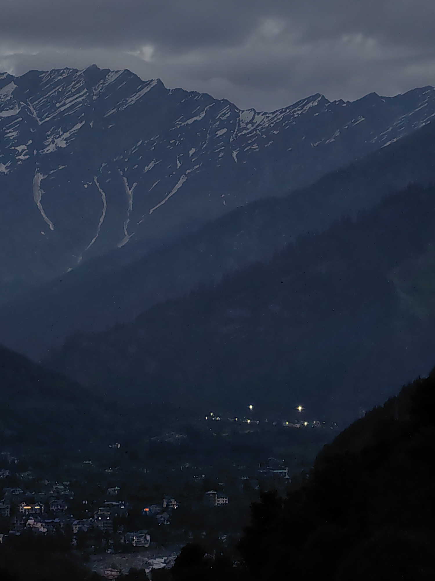 The image depicts a wide, mountainous landscape at dusk or dawn. In the foreground, the dark silhouette of a tree-covered slope dominates the lower right portion of the frame. Beyond this, a valley opens up, revealing a settlement dotted with illuminated buildings, suggesting the town of Vashisht, India. The lights from the town cast a faint glow. Further in the background, layers of darker, forested mountains recede into the distance. The uppermost layer of the scene features a majestic mountain range with snow-capped peaks, their rugged textures softened by the dim light and atmospheric haze. The sky above is overcast with dark, heavy clouds, contributing to the overall moody and subdued lighting of the scene. There are no people or discernible actions visible.