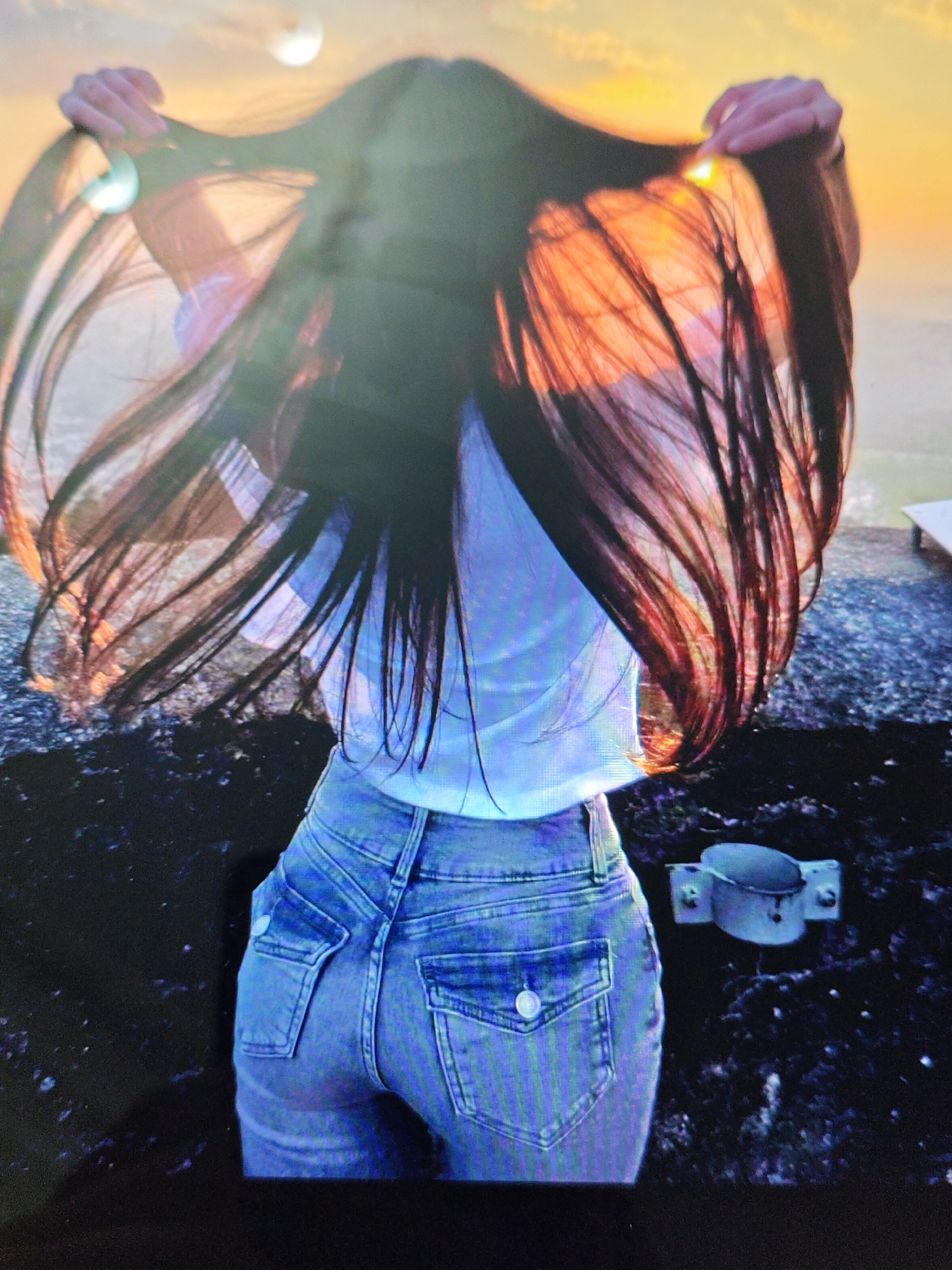 A woman stands with her back to the camera, holding her long dark hair up and out, framing her face which is obscured by her hair. The sun is setting, casting a warm orange glow through her hair and across the sky. She is wearing a light-colored t-shirt and blue jeans. The ground beneath her appears to be a dark, pebbly surface. In the lower right quadrant of the image, a metal clamp is attached to a post. The setting appears to be outdoors, possibly near the Caspian Sea in Baku, Azerbaijan, given the atmospheric conditions and the potential for a coastal view.