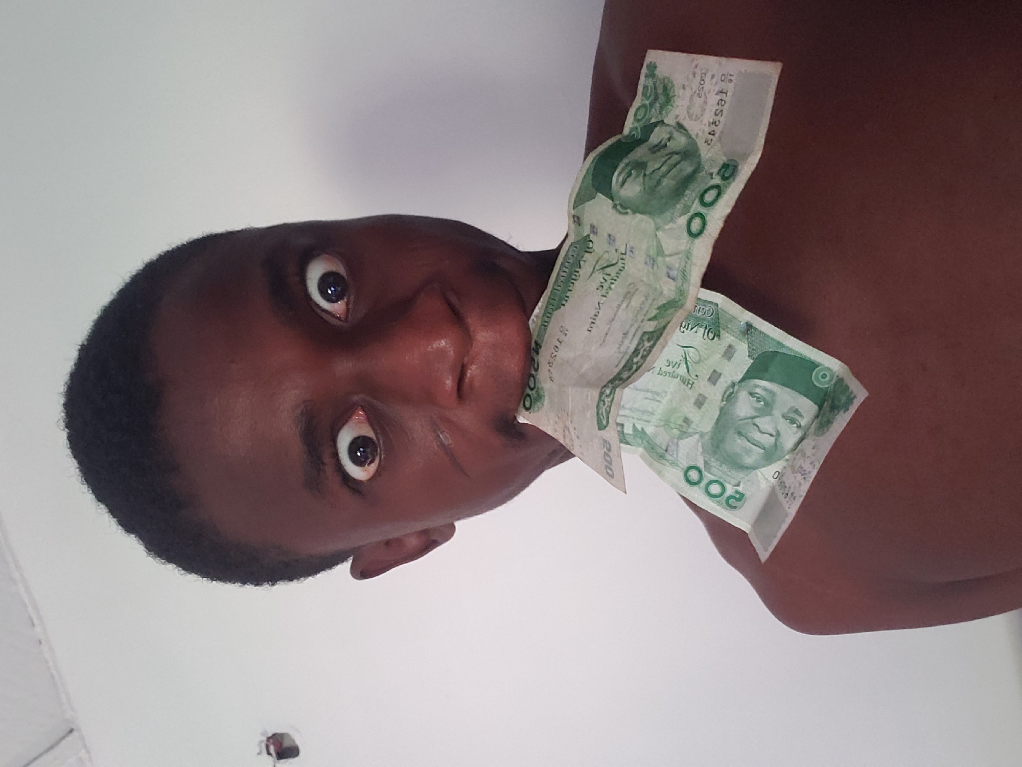 A young person, likely male, with dark skin and short, dark hair, is the central subject of this close-up, angled photograph. Their head and upper torso are visible against a plain, light-colored background, possibly a wall or ceiling, suggesting an indoor setting in Adonkolo, Nigeria.
The individual is posing with an exaggerated expression, featuring wide, bulging eyes and a slightly open mouth, conveying a sense of surprise, excitement, or a playful and comical demeanor. Two light green Nigerian 200 Naira banknotes are prominently displayed on their upper chest and shoulder area. One note features the portrait of Alhaji Aliyu Mai-Bornu in a green traditional cap, while the other side, partially visible, depicts the Central Bank of Nigeria headquarters building.
Visible text on the banknotes includes "CENTRAL BANK OF NIGERIA", the denomination "200", and "TWO HUNDRED NAIRA". Partial serial numbers such as "BDS2 F65392" and "DS2 T65392" can also be observed. The image is captured from a low angle, making it appear as though the subject is looking down, or the camera is positioned below them. The lighting is bright and even, without any specific cues for the time of day or weather. A small, dark fixture is visible on the background surface in the lower left corner.