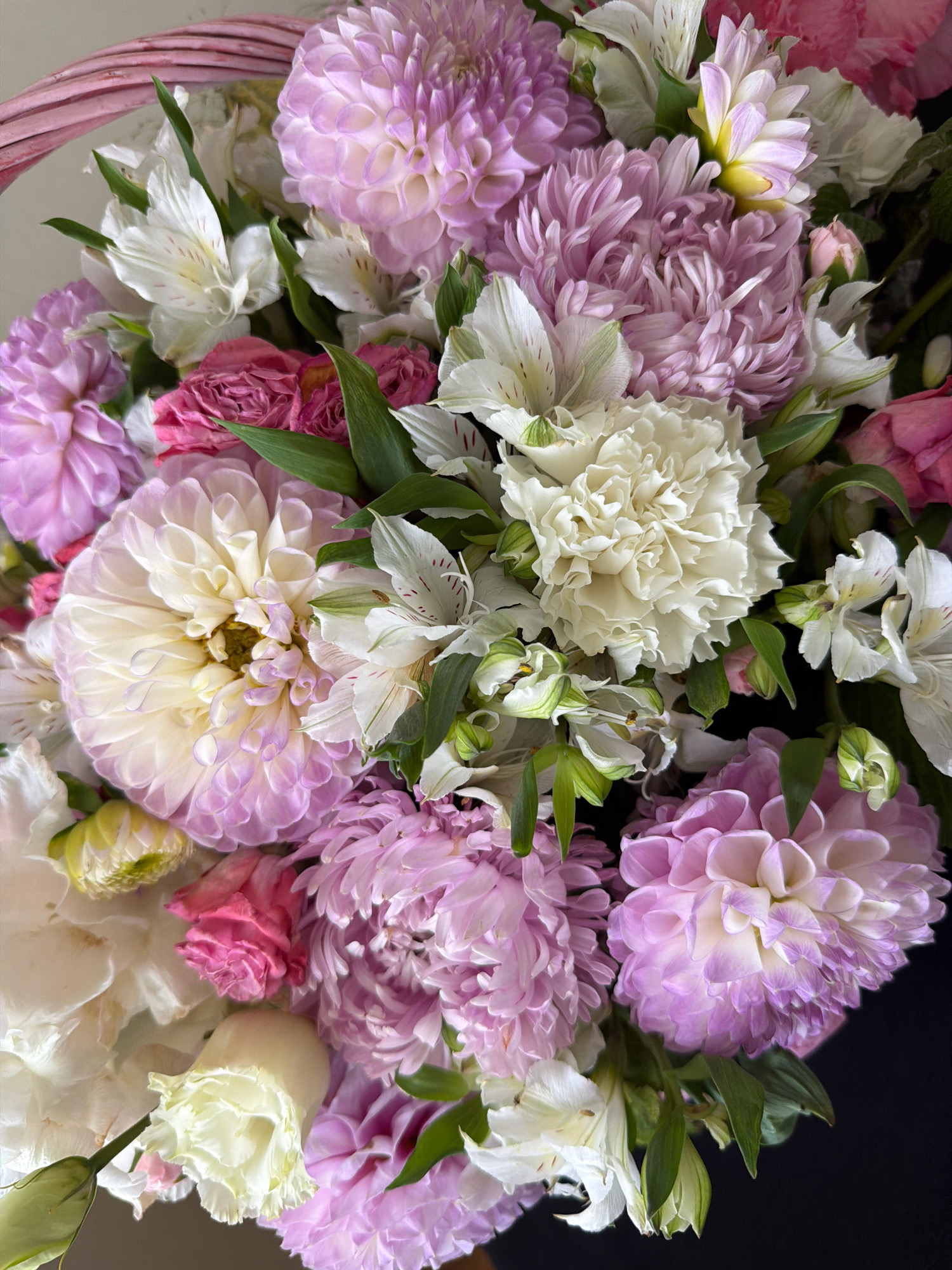 This is a close-up, overhead shot of a lush bouquet of flowers. The bouquet is predominantly composed of large, ruffled dahlias in shades of pale pink, lavender, and cream. Interspersed among the dahlias are delicate white alstroemeria flowers with subtle pink markings. There are also a few small, deep pink rosebuds. The flowers are arranged densely, with vibrant green foliage peeking through. The background is dark, with a hint of a pink woven basket at the top left corner. There are no people or actions depicted in the image, and the setting is not identifiable as being in Бабініцкі сельскі Савет, Belarus.