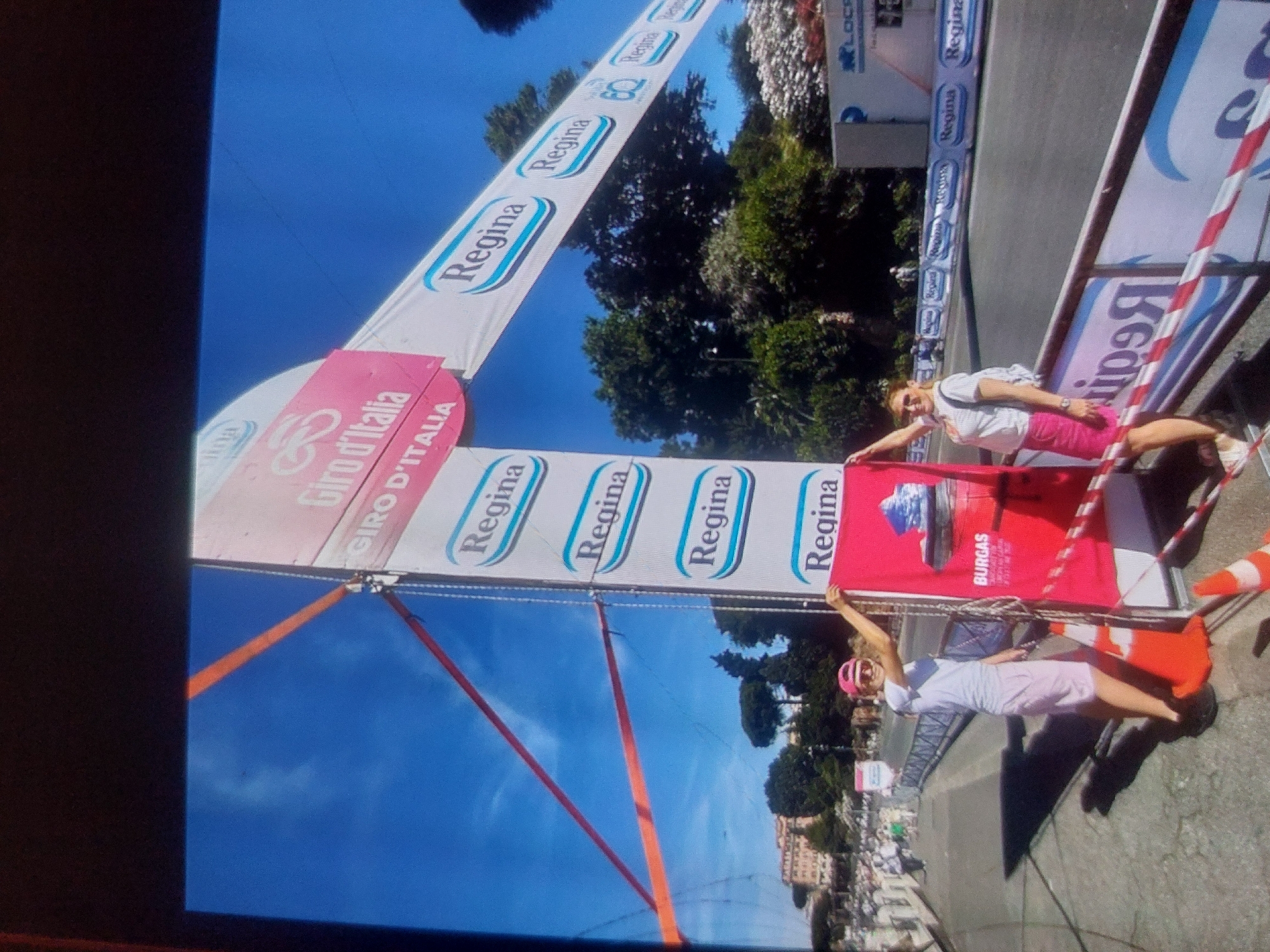 Here is a description of the image:
The image shows what appears to be a finish line banner for the "Giro d'Italia" race, prominently featuring the name "Regina" repeated across the banner. The location appears to be in an urban area, given the presence of buildings and a street with fencing or barriers along the sides. There are two women in the image near the banner. One woman is wearing a white shirt and pink pants, while the other woman is wearing a white dress and a pink helmet. It appears the women are interacting with the banner as they pose for the image. In the foreground, traffic cones are present. There are no indications to confirm this is the city of Tsarevo, Bulgaria.